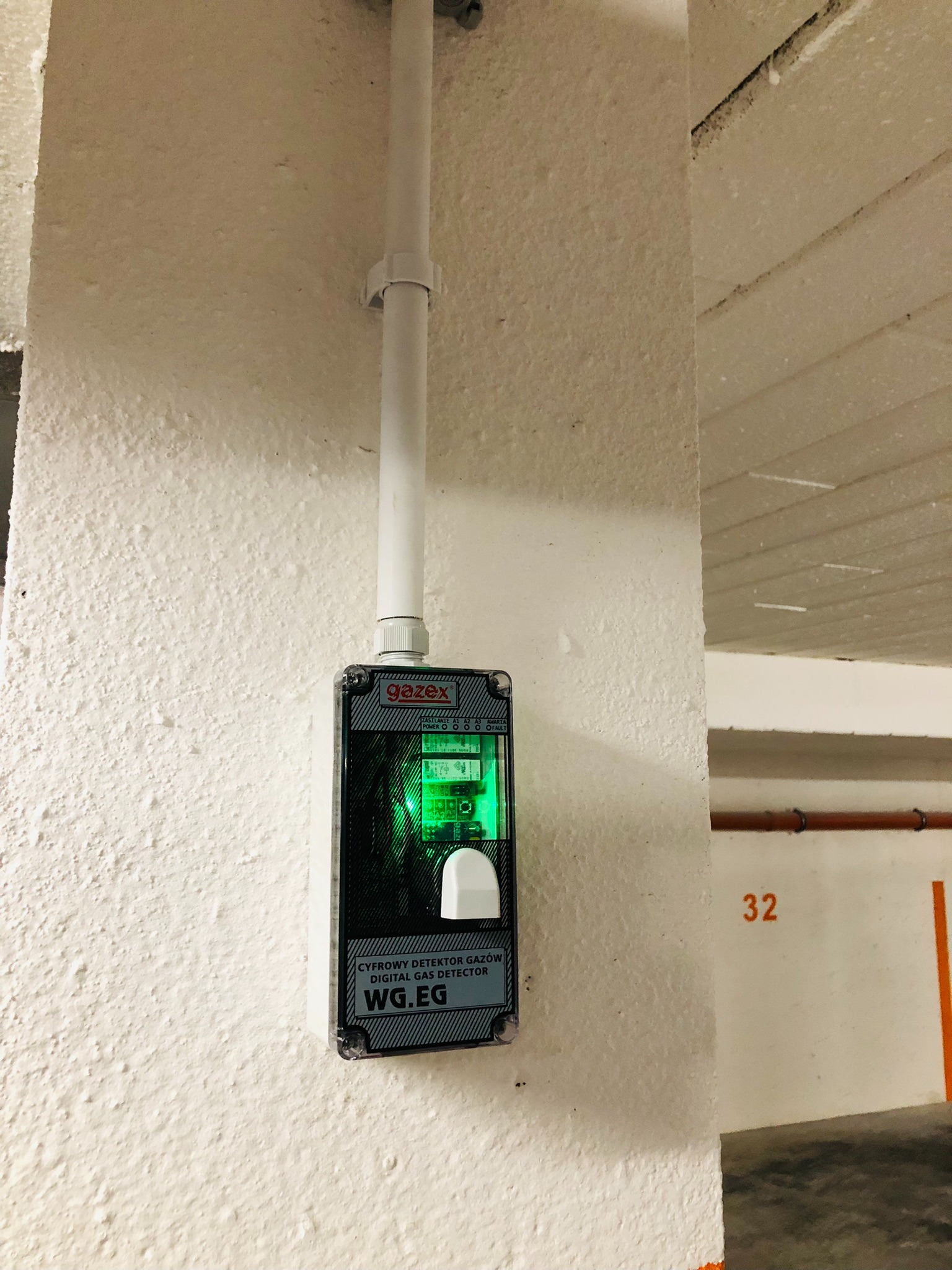 Cyfrowy detektor gazów WG.EG marki Gazex zamontowany na pomalowanej na biało ścianie w garażu podziemnym, z widoczną rurką instalacyjną powyżej.