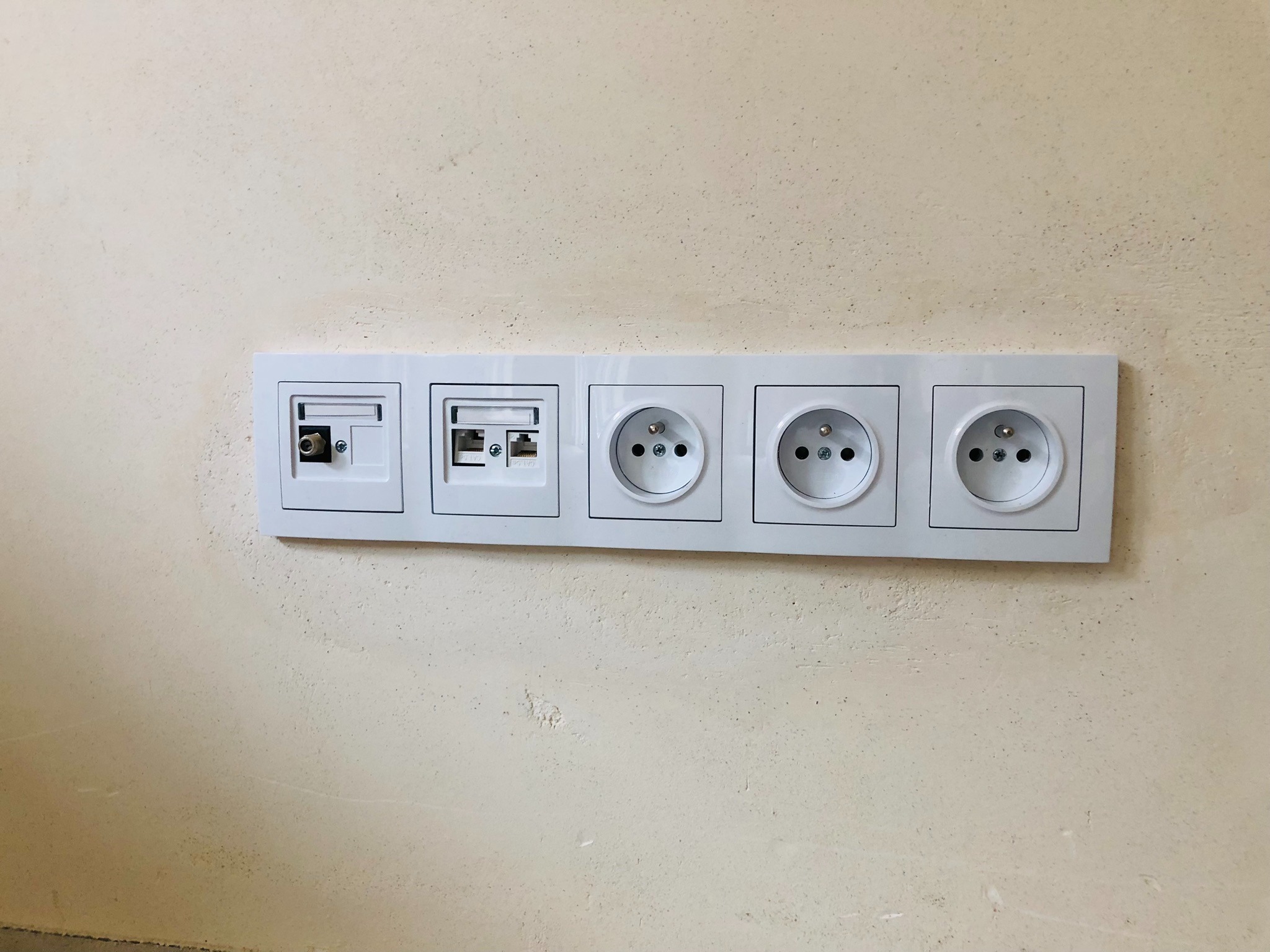 Panel ścienny z gniazdami: antenowe, internetowe RJ45 i trzy gniazdka elektryczne, zamontowany na surowej ścianie.