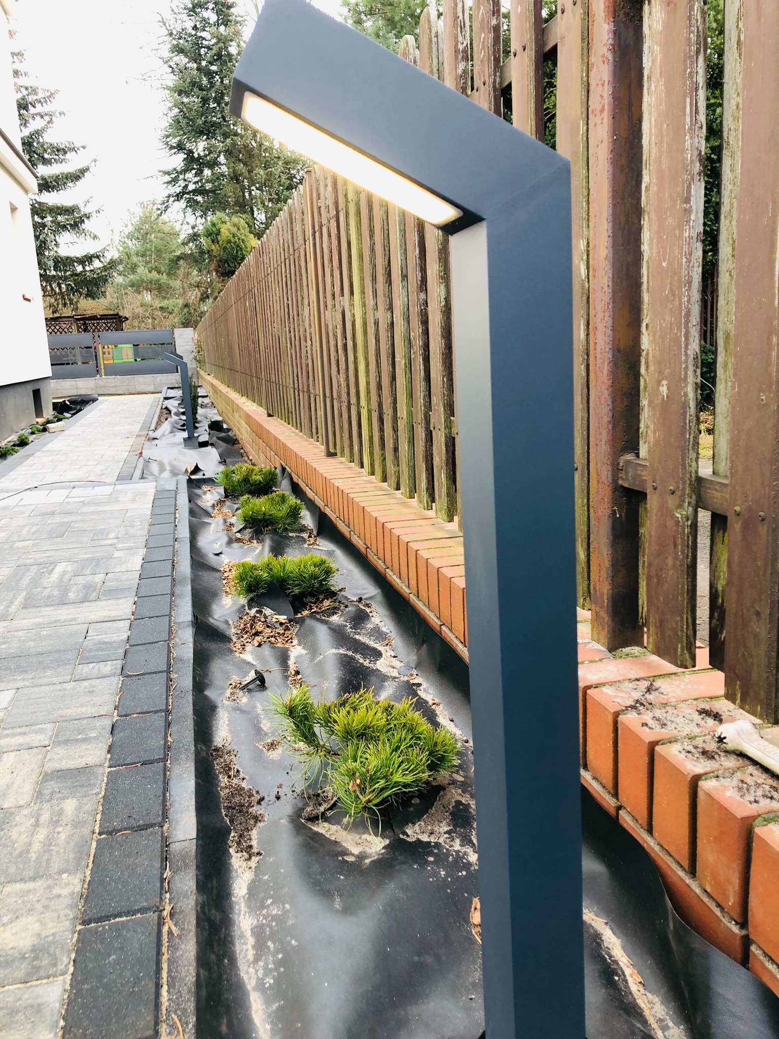 Nowoczesna, grafitowa lampa ogrodowa LED o geometrycznym kształcie, zainstalowana w Wieliszewie wzdłuż ścieżki i drewnianego ogrodzenia z ceglaną podmurówką, oświetlająca nowo posadzone rośliny.