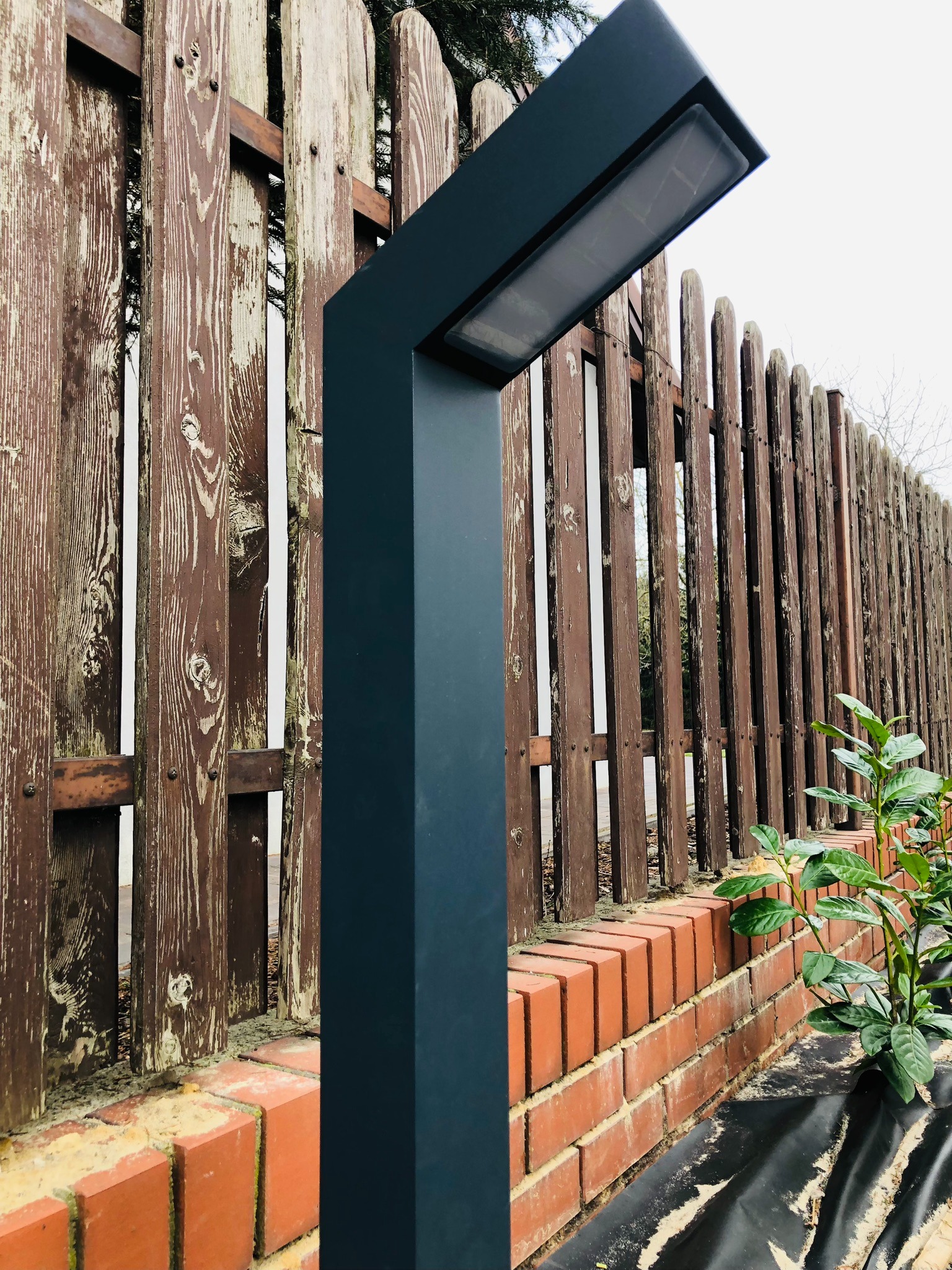 Nowoczesna lampa ogrodowa w kolorze grafitowym, oświetlająca drewniany płot z ceglaną podmurówką i młodymi roślinami.