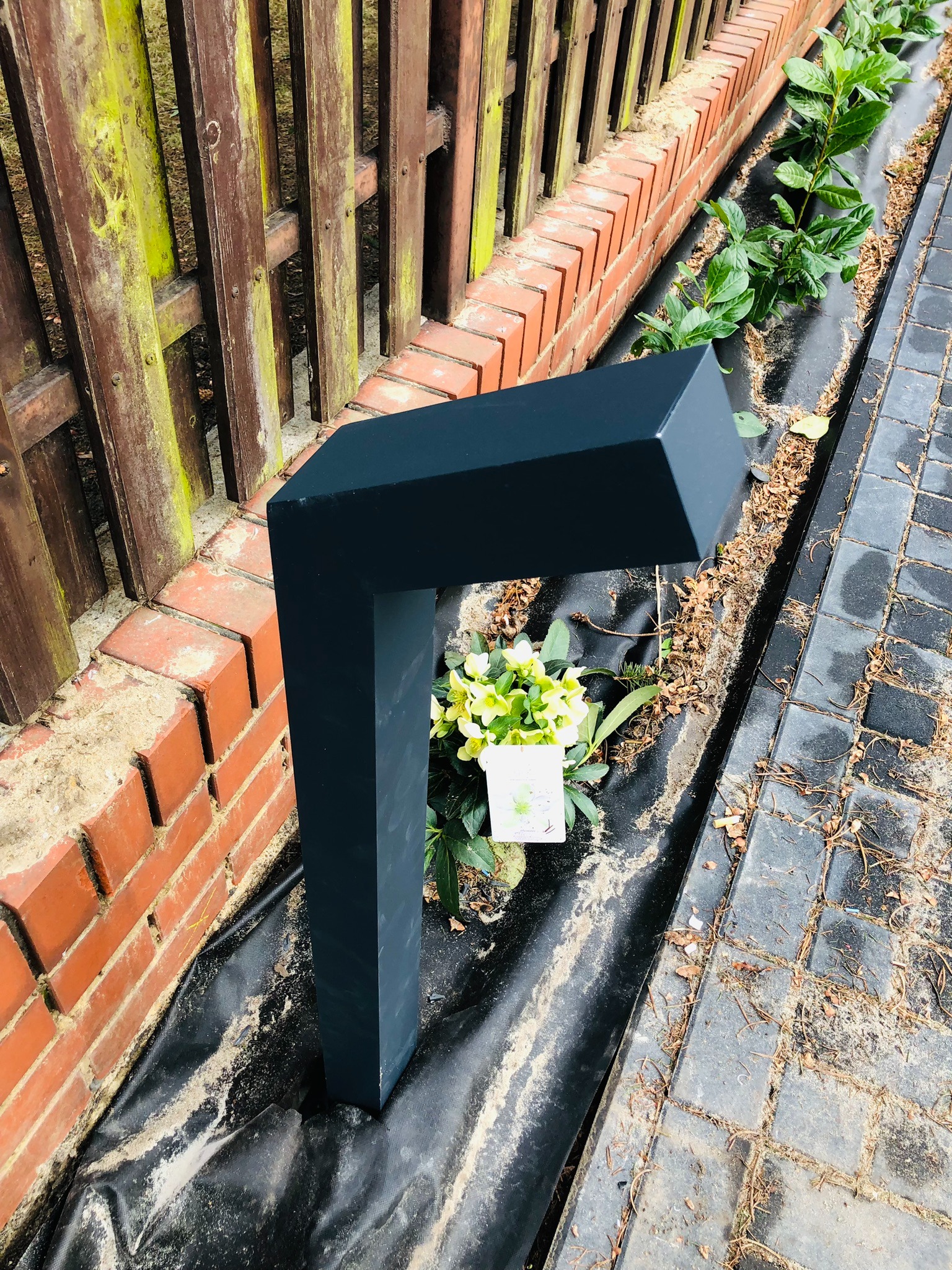 Nowoczesna, grafitowa lampa ogrodowa w kształcie litery L, umieszczona wzdłuż murku z cegły i drewnianego płotu, otoczona roślinami i wyściółką.