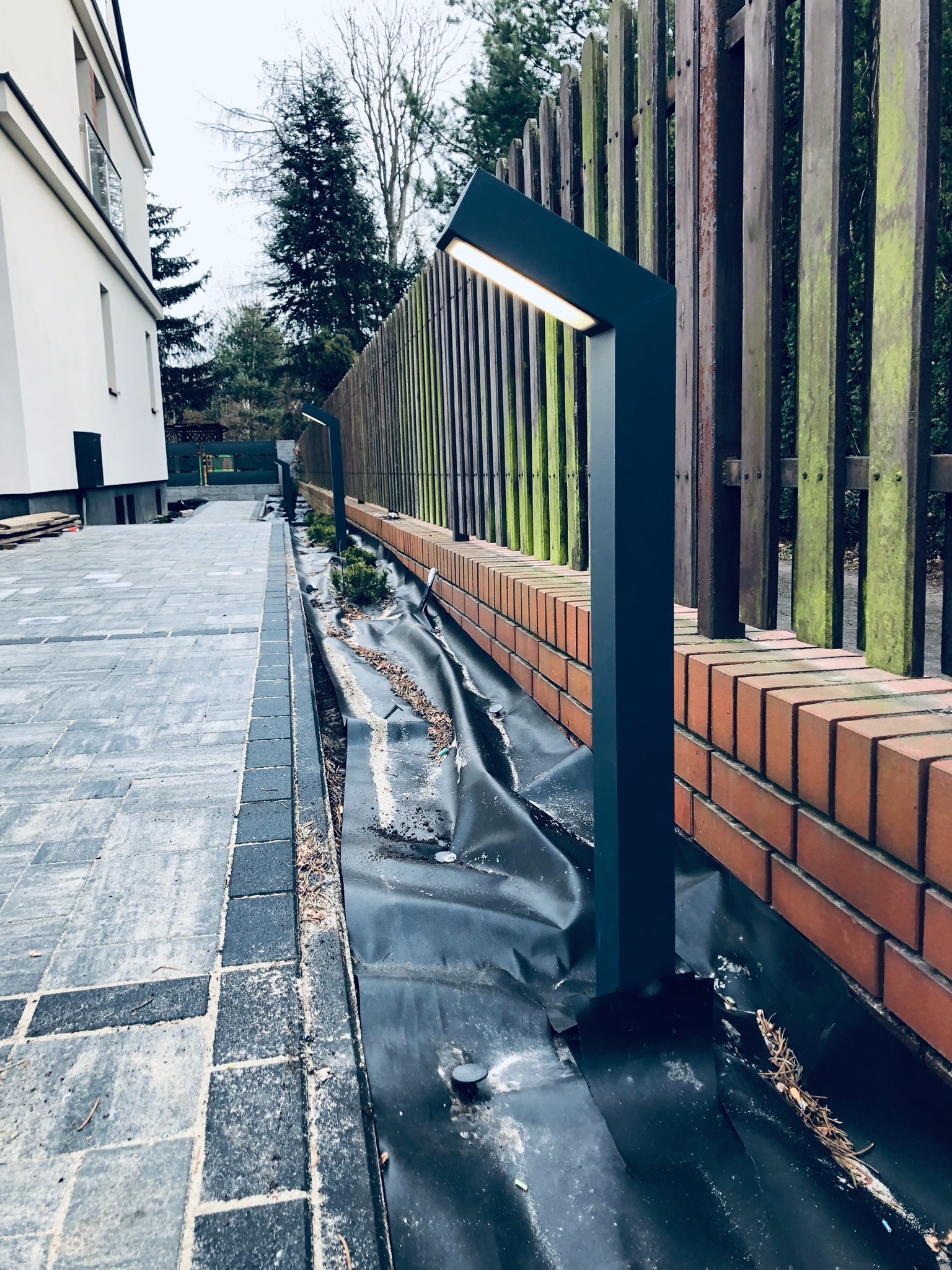 Nowoczesna, antracytowa lampa ogrodowa LED o geometrycznym kształcie, oświetlająca chodnik z kostki brukowej i drewniany płot z mchem, na tle białego budynku.