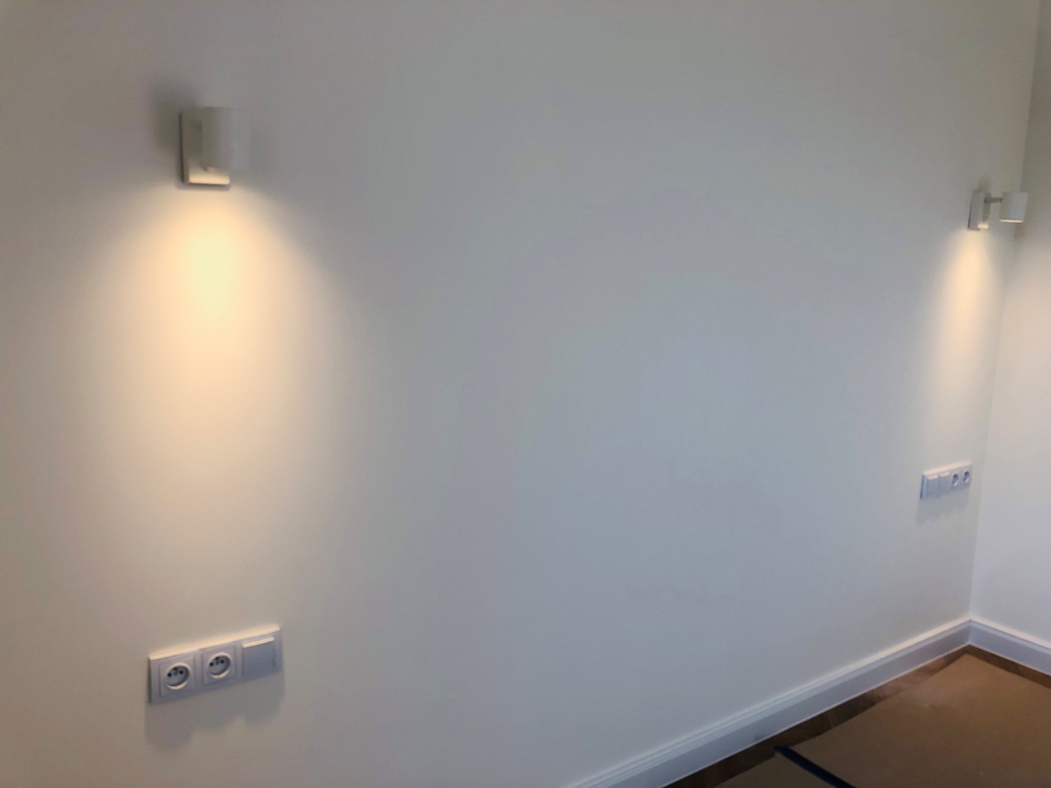 Dwa nowoczesne, kwadratowe kinkiety ścienne, każdy z ciepłym, stożkowym strumieniem światła, umieszczone na gładkiej, białej ścianie z zamontowanymi podwójnymi gniazdkami elektrycznymi...