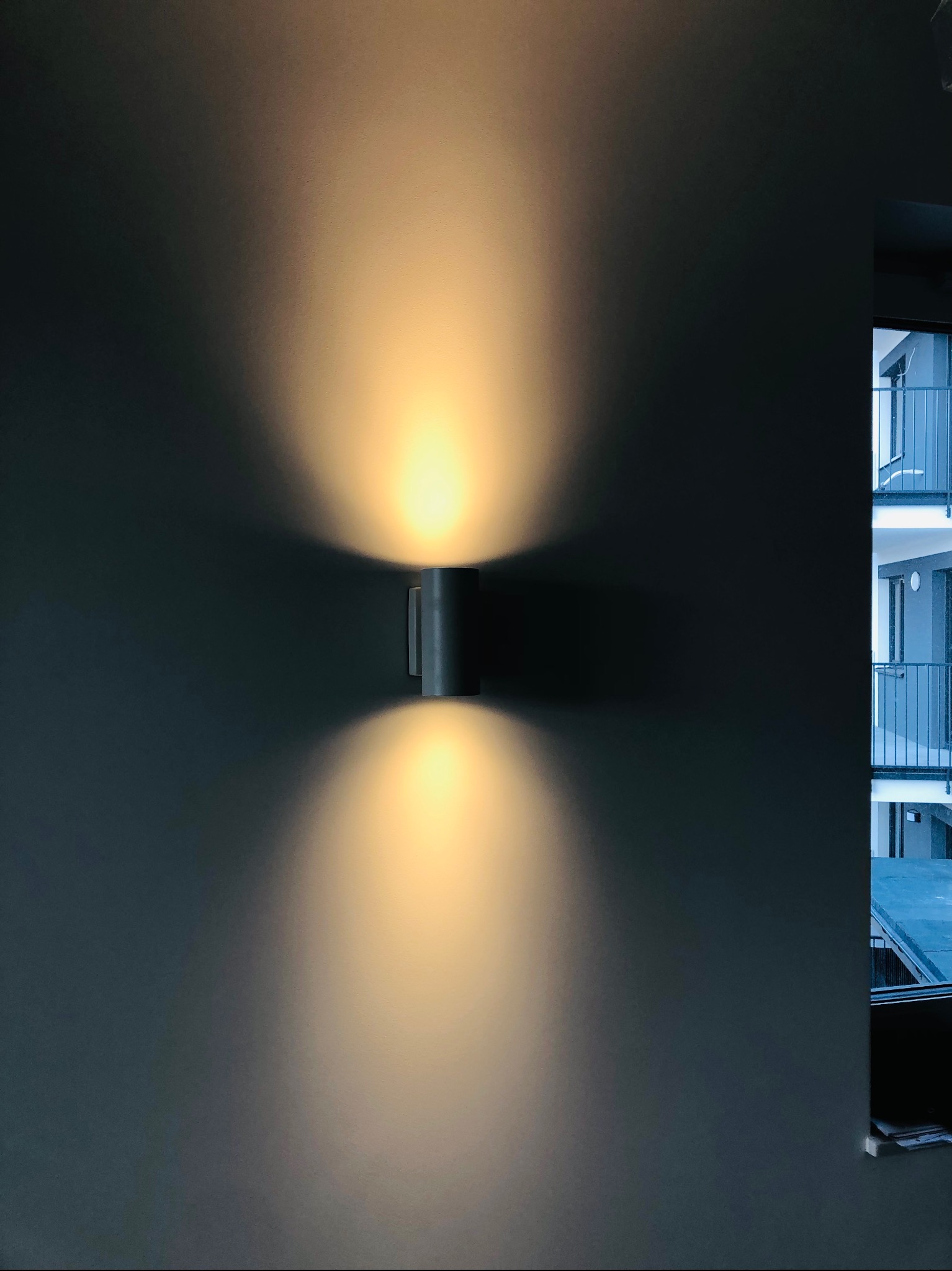Świecąca, cylindryczna lampa ścienna, emitująca światło w górę i w dół, zamontowana na gładkiej, szarej ścianie w nowoczesnym wnętrzu, z widokiem na balkony sąsiedniego budynku przez okno.