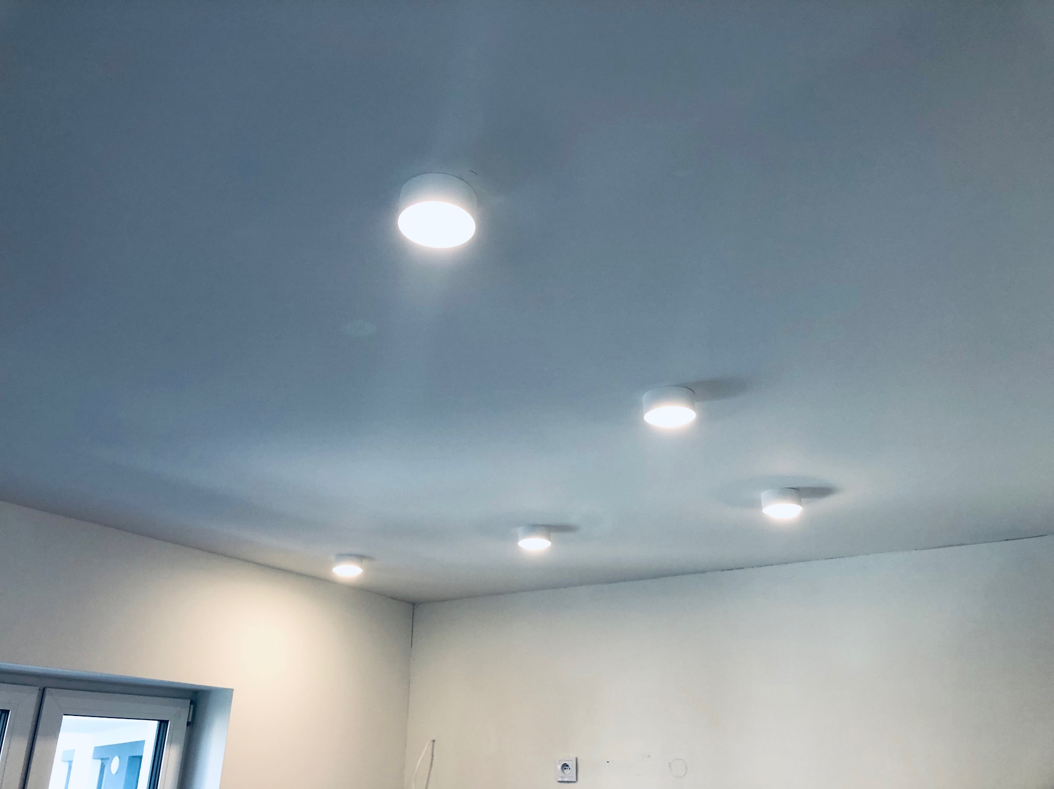 Biały sufit z zamontowanymi sześcioma okrągłymi lampami LED, widok z dołu na wnętrze w trakcie wykańczania, ściany pomalowane na biało, widoczne gniazdko elektryczne i fragment okna.