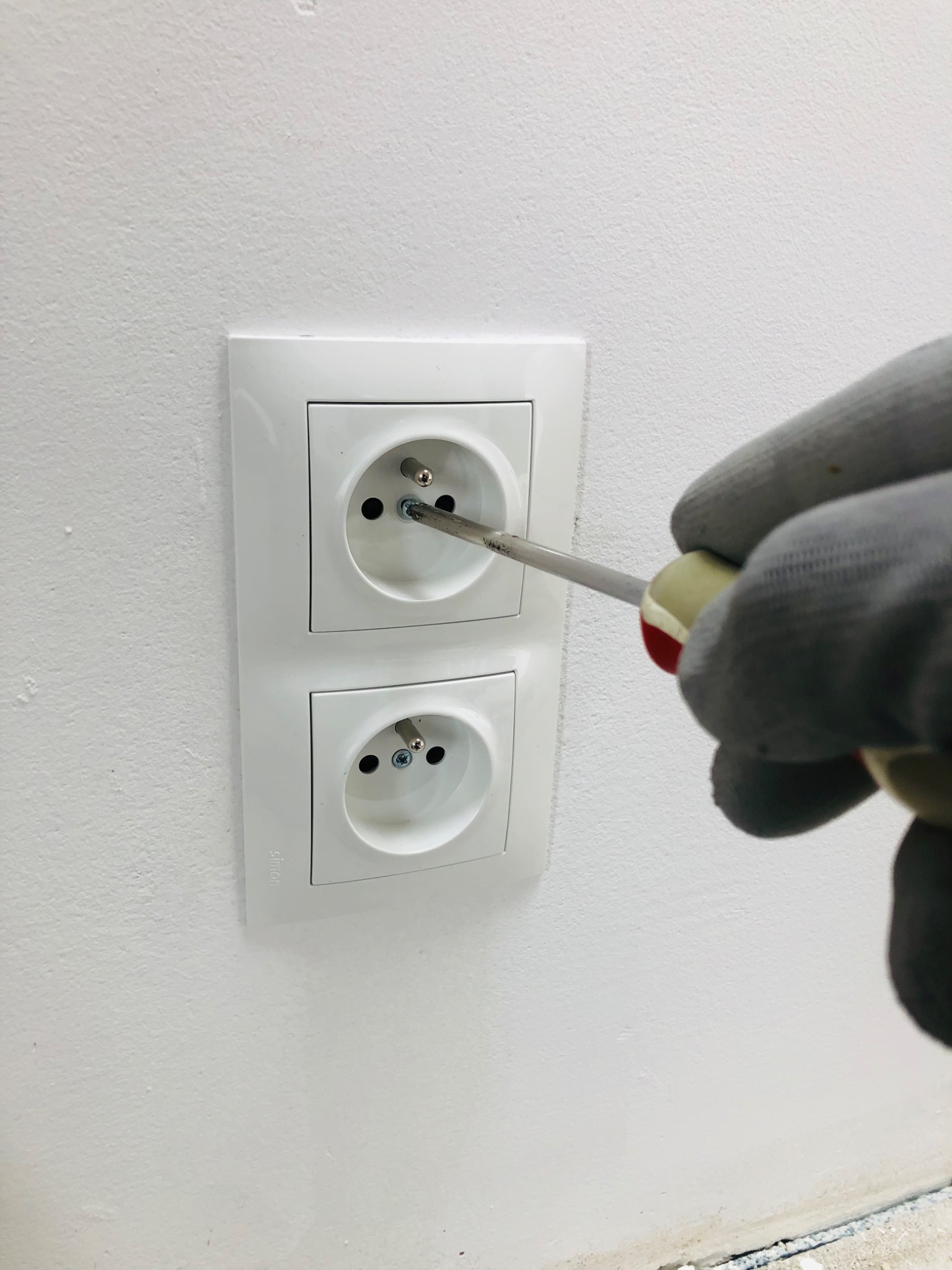 Elektryk dokręca śrubę w gniazdku elektrycznym za pomocą śrubokręta. Biała ściana w tle. Widoczne dwa gniazdka.
