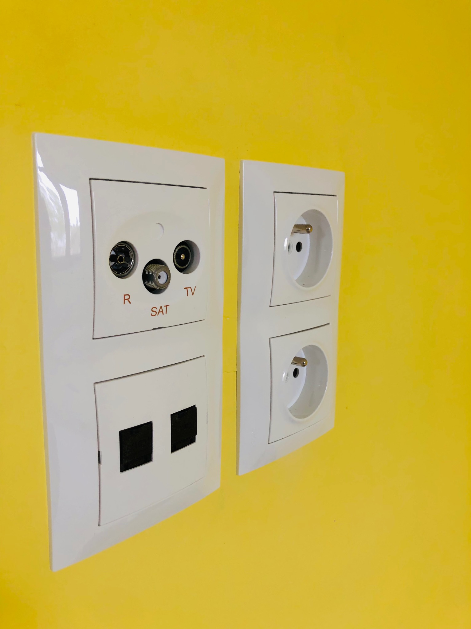 Białe gniazdka elektryczne i antenowe (R, SAT, TV) na jaskrawożółtej ścianie, zamontowane równolegle. Dwa gniazdka z bolcem uziemiającym.