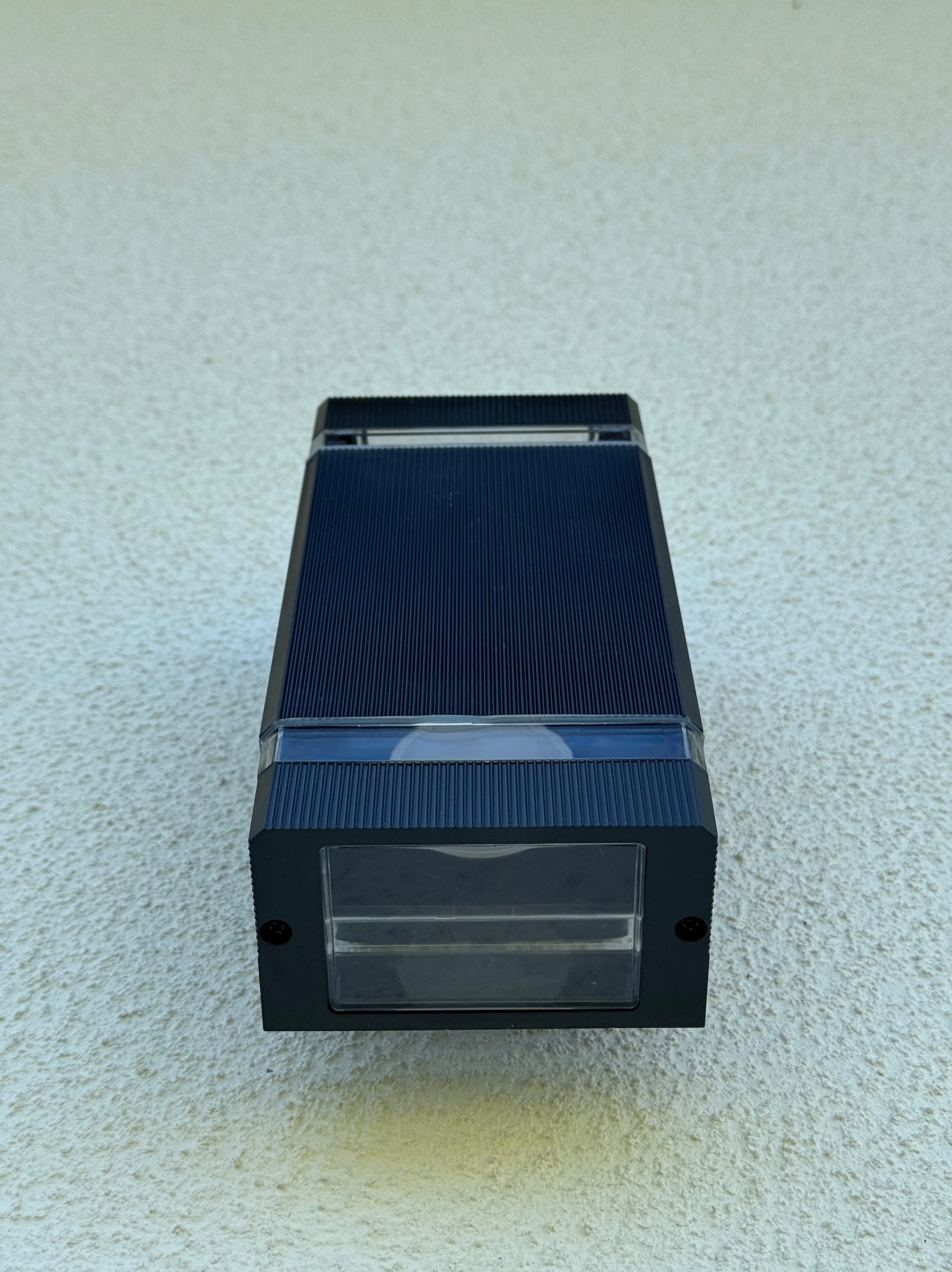 Zamontowany, nowoczesny kinkiet elewacyjny z czarną obudową i przezroczystym kloszem, widoczny na tle jasnej, teksturowanej ściany zewnętrznej.