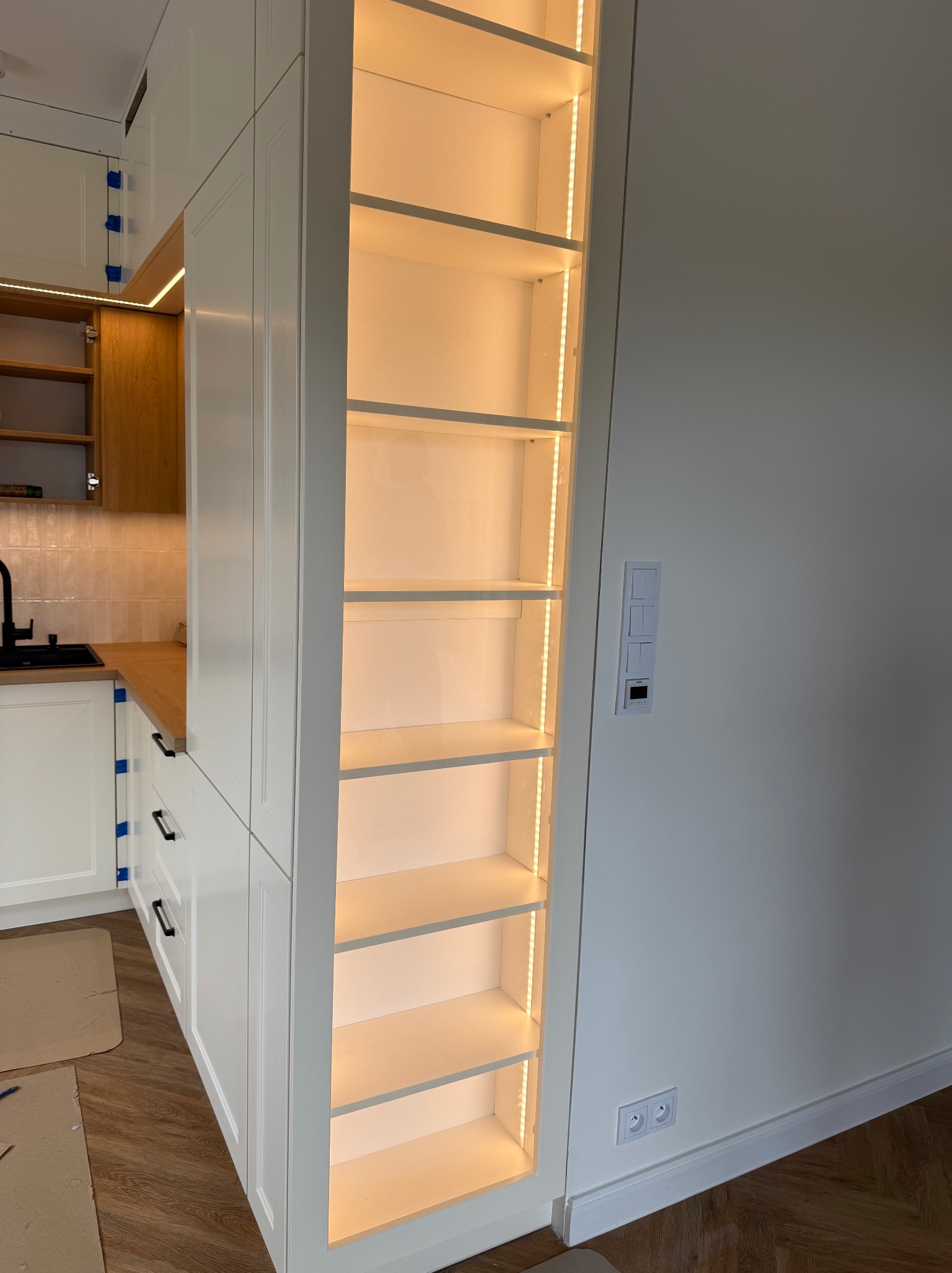 Biała zabudowa kuchenna z półkami podświetlonymi ciepłym światłem LED. Widoczne gniazdka i włączniki na ścianie. Parkiet na podłodze.