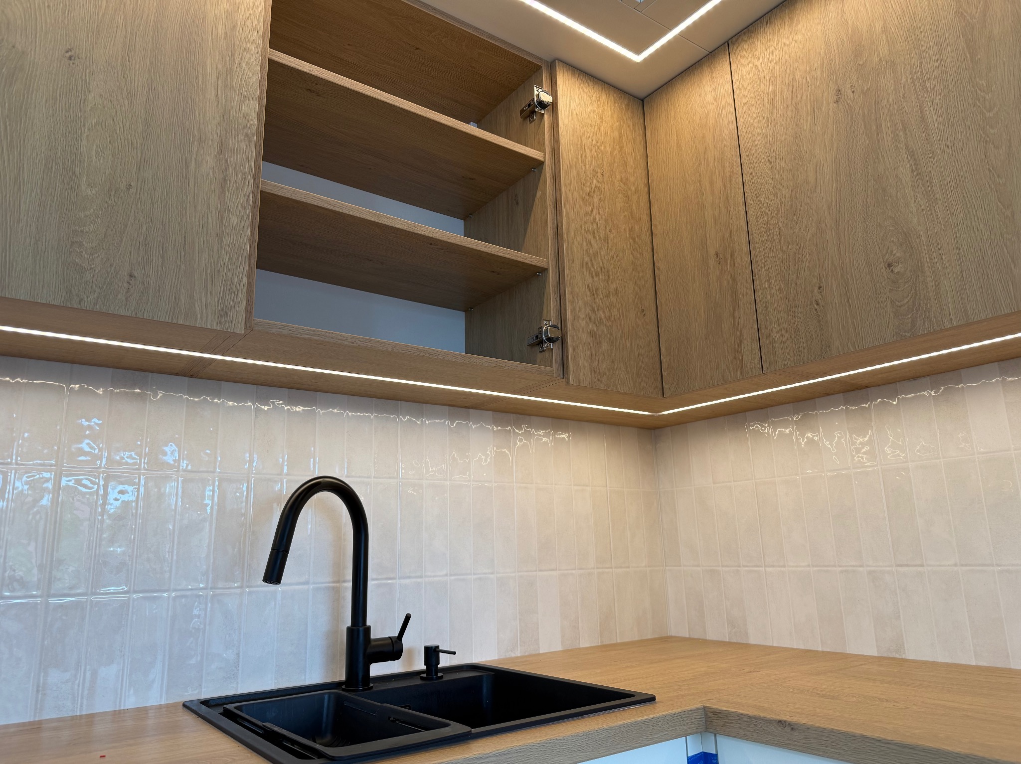 Kuchnia z drewnianymi szafkami, czarnym zlewem i baterią. Oświetlenie LED podszafkowe i sufitowe. Otwarta szafka eksponuje półki.