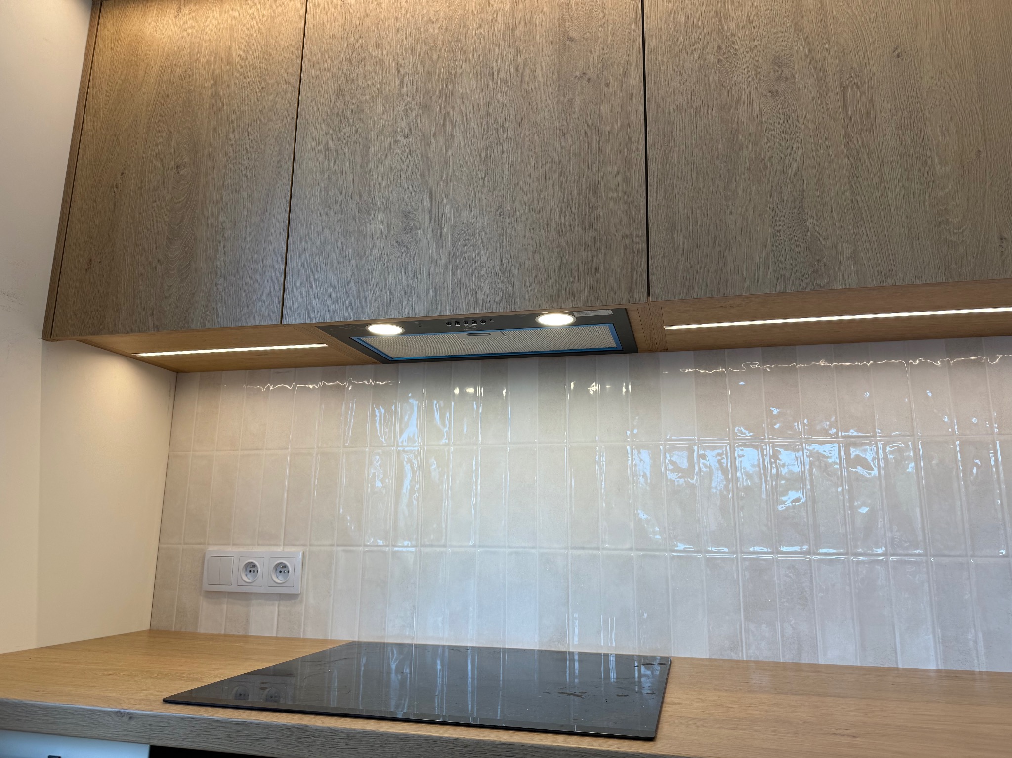 Kuchnia z drewnianymi szafkami, okapem i oświetleniem LED pod szafkami. Płyta indukcyjna na blacie. Białe, błyszczące płytki na ścianie i gniazdka elektryczne.