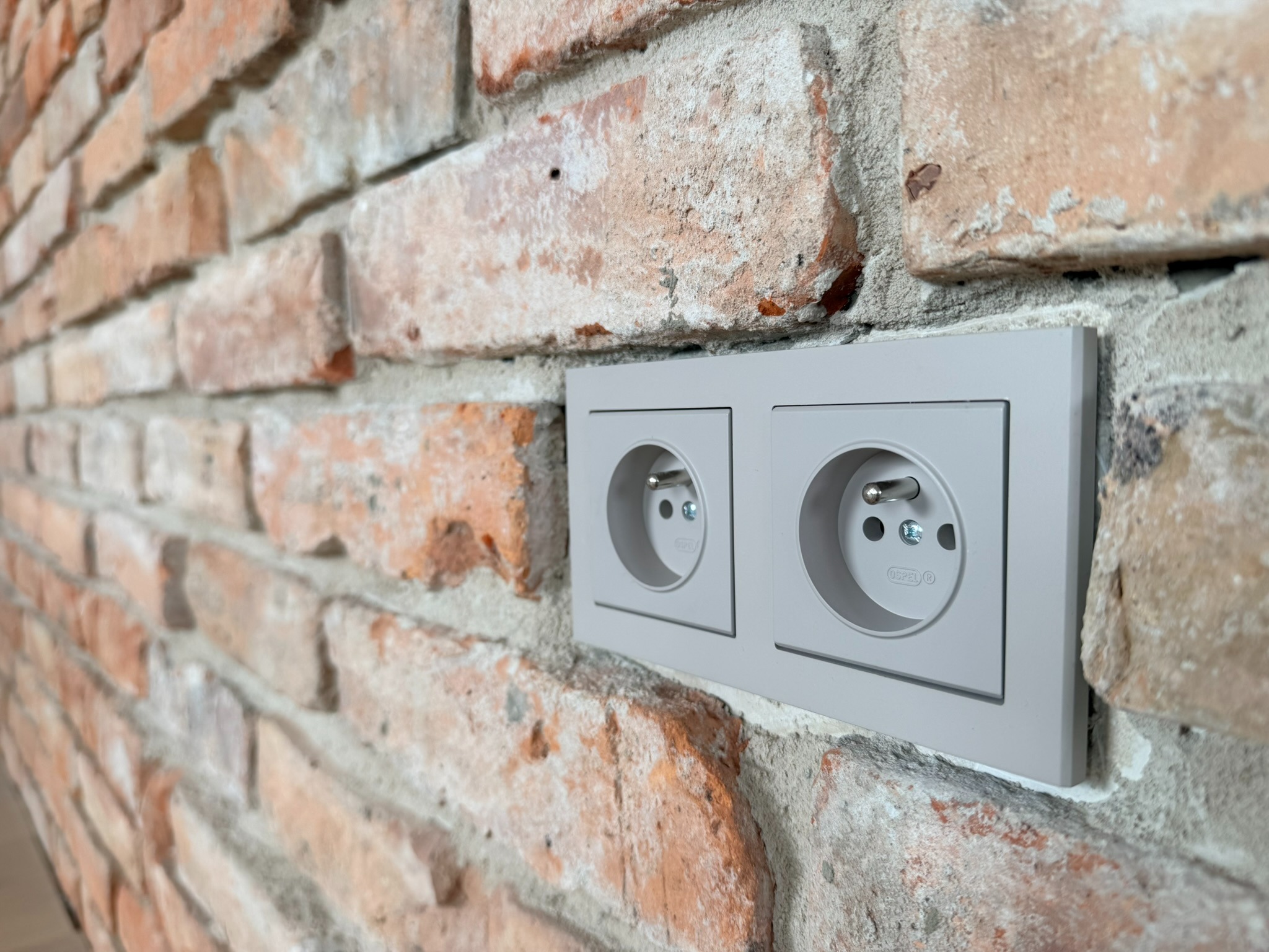 Dwa gniazdka elektryczne w szarej ramce, zamontowane na ścianie z czerwonej cegły o nieregularnej fakturze. Widok z bliska, z boku.