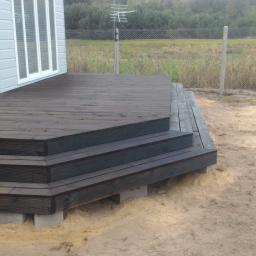 Multimo Buduj Remontuj - Drewniany taras z trzema schodami, świeżo po impregnacji, wsparty na betonowych bloczkach, obok stoi mężczyzna w roboczych spodniach na tle ogrodzenia z siatki i zieleni.