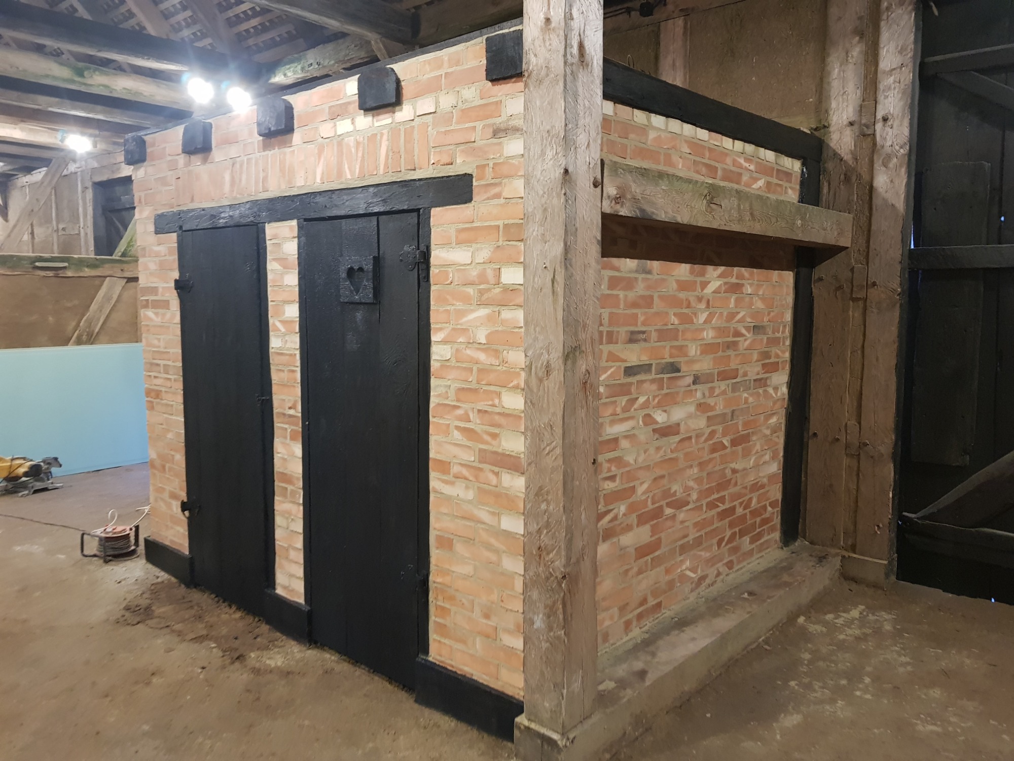 Murowana konstrukcja z cegły z czarnymi, drewnianymi drzwiami z wyciętym sercem, wsparta na drewnianych belkach wewnątrz budynku gospodarczego.