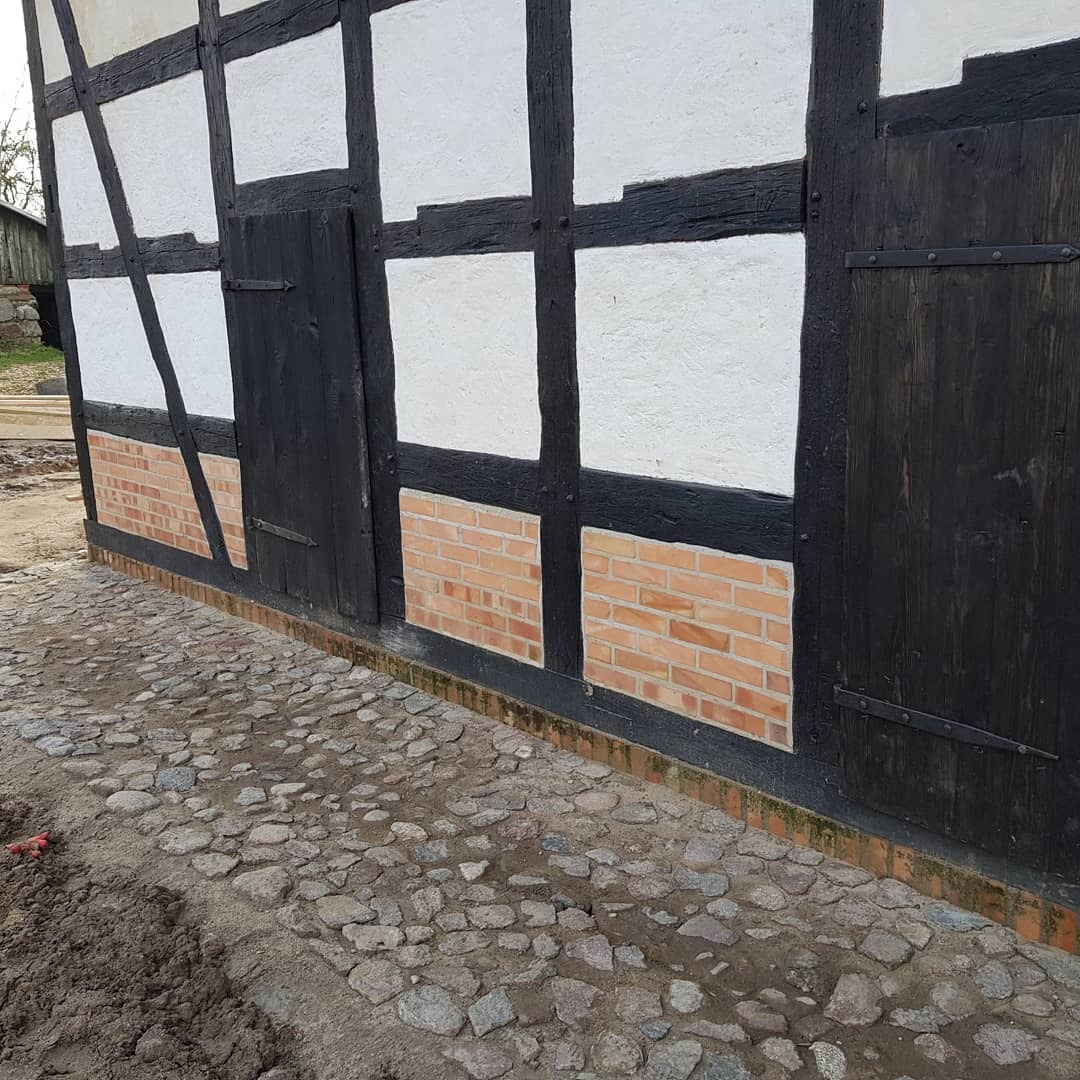 Fragment ściany szachulcowej, z widocznym murem z cegły klinkierowej w dolnej części i brukowaną nawierzchnią z kamienia polnego.