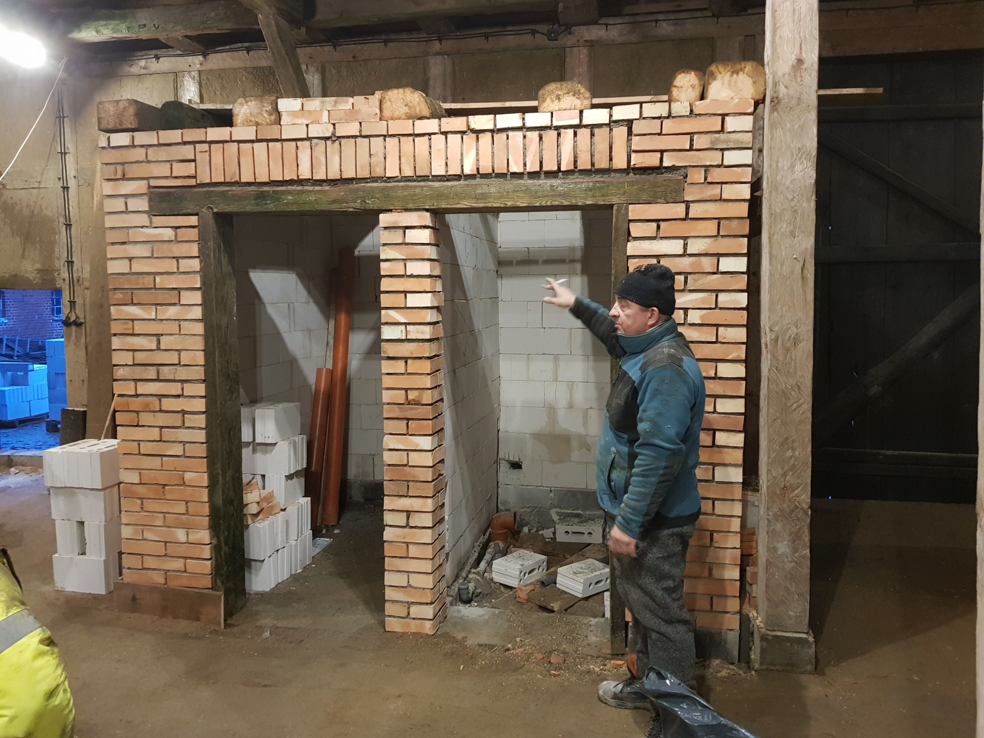Wznoszenie ścianek działowych z cegły w surowym wnętrzu budynku, widoczne otwory drzwiowe z drewnianymi nadprożami i stropy z belek, mężczyzna w roboczym ubraniu wskazuje na konstrukcję.