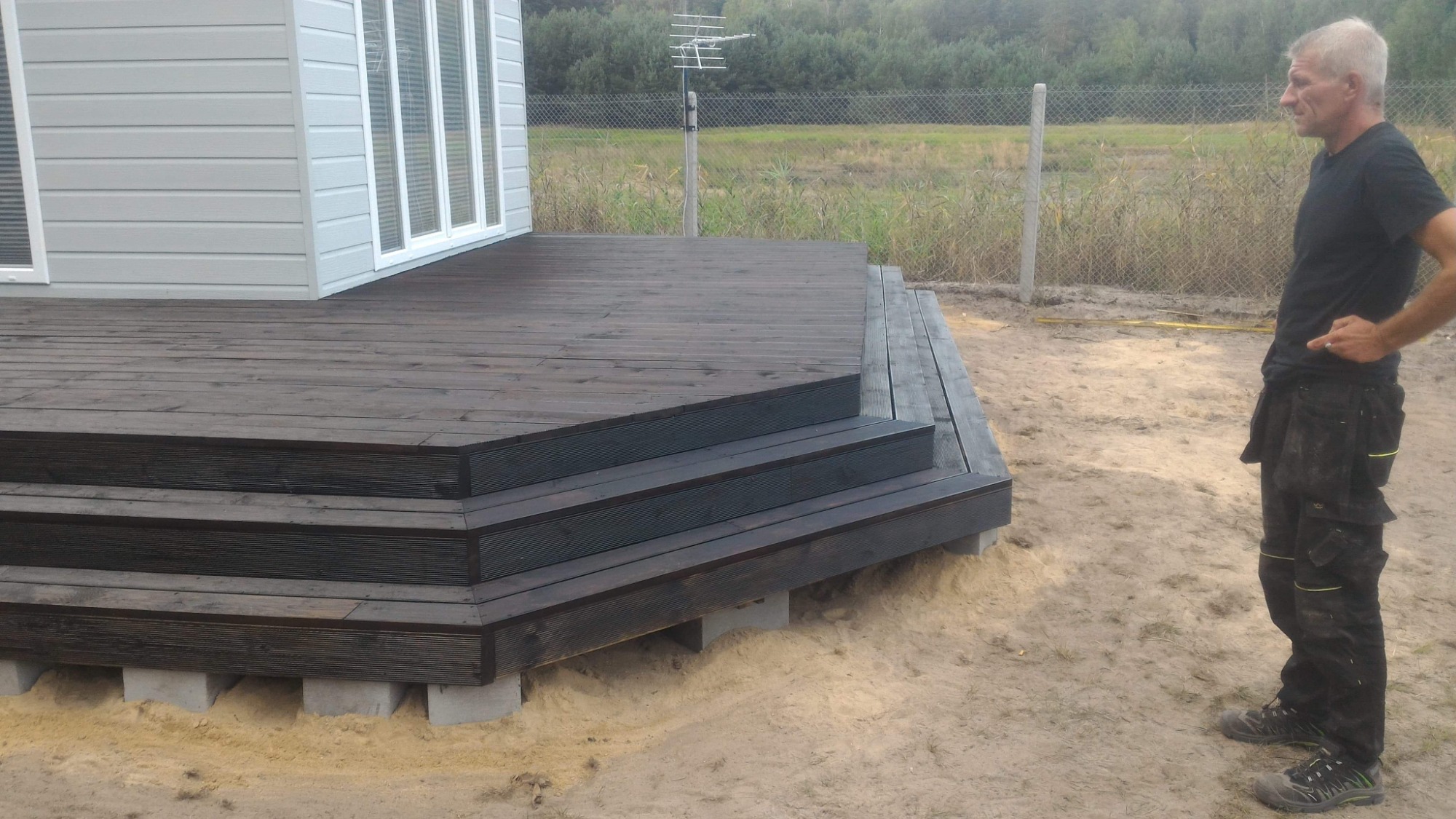 Drewniany taras z trzema schodami, świeżo po impregnacji, wsparty na betonowych bloczkach, obok stoi mężczyzna w roboczych spodniach na tle ogrodzenia z siatki i zieleni.