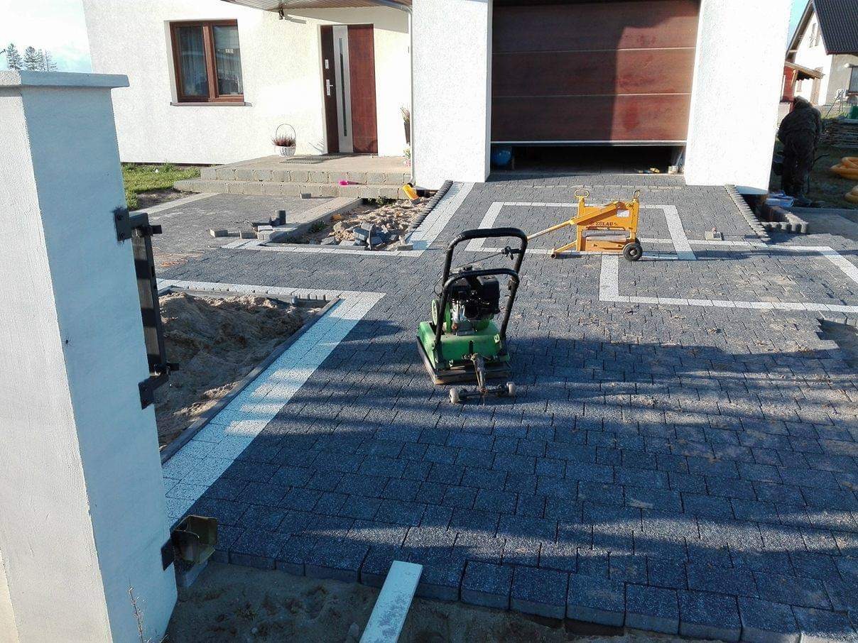 Układanie ciemnoszarej kostki brukowej z białymi akcentami na podjeździe przed domem; widoczna zagęszczarka w trakcie pracy i żółty podnośnik do kostki.