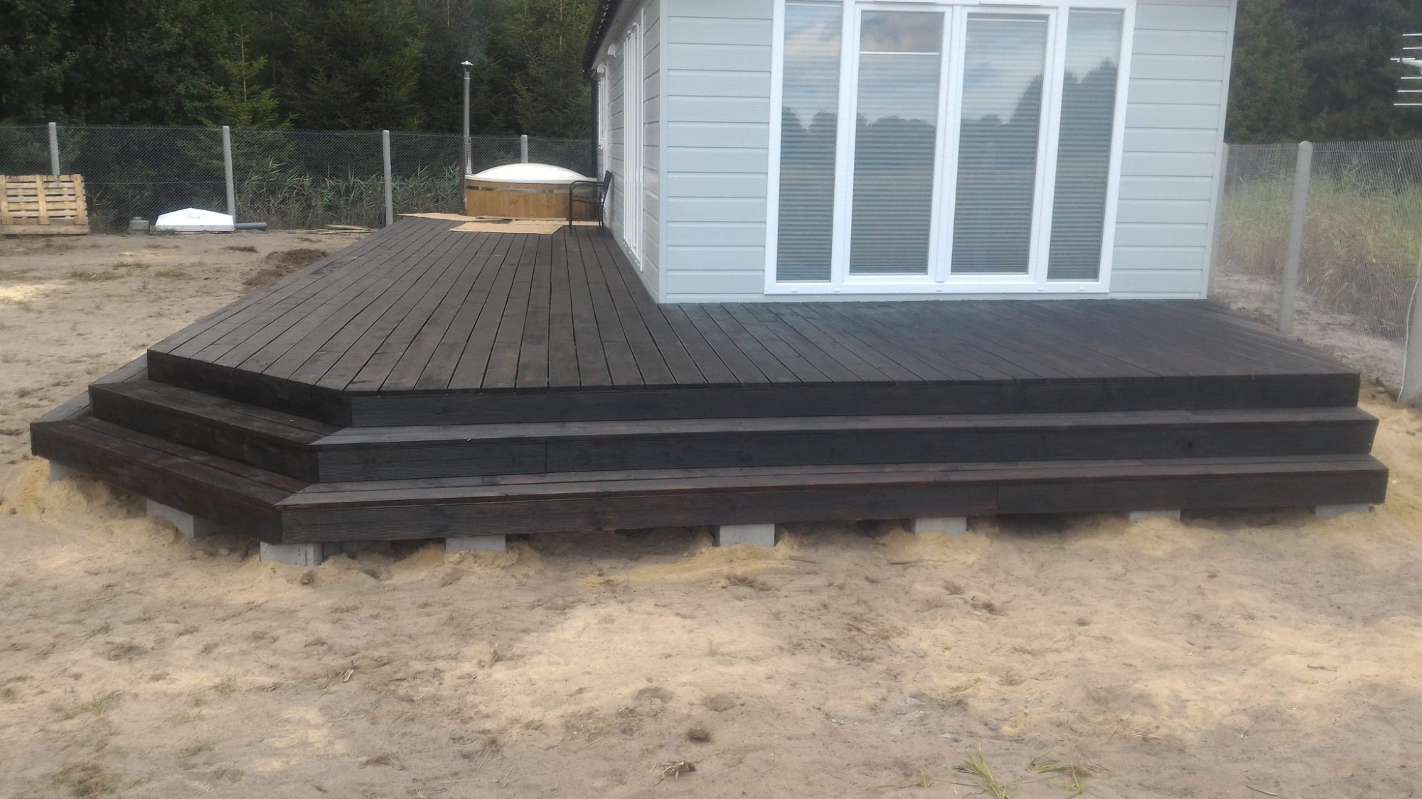 Drewniany taras z trzema stopniami, pomalowany na ciemny brąz, przylegający do szarego domku z białymi oknami, widoczny fragment jacuzzi i palety w tle.