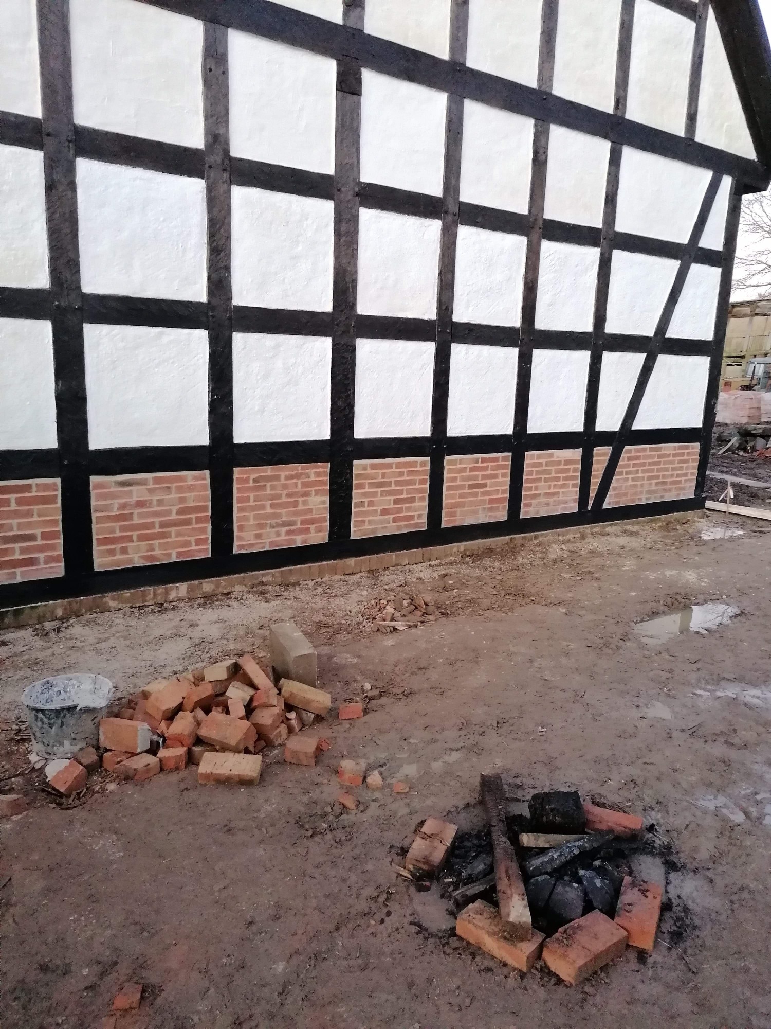 Ściana budynku w konstrukcji szkieletowej z wypełnieniem muru pruskiego, częściowo obłożona cegłą, z widocznymi stertami cegieł i pozostałościami po ognisku na ziemi.