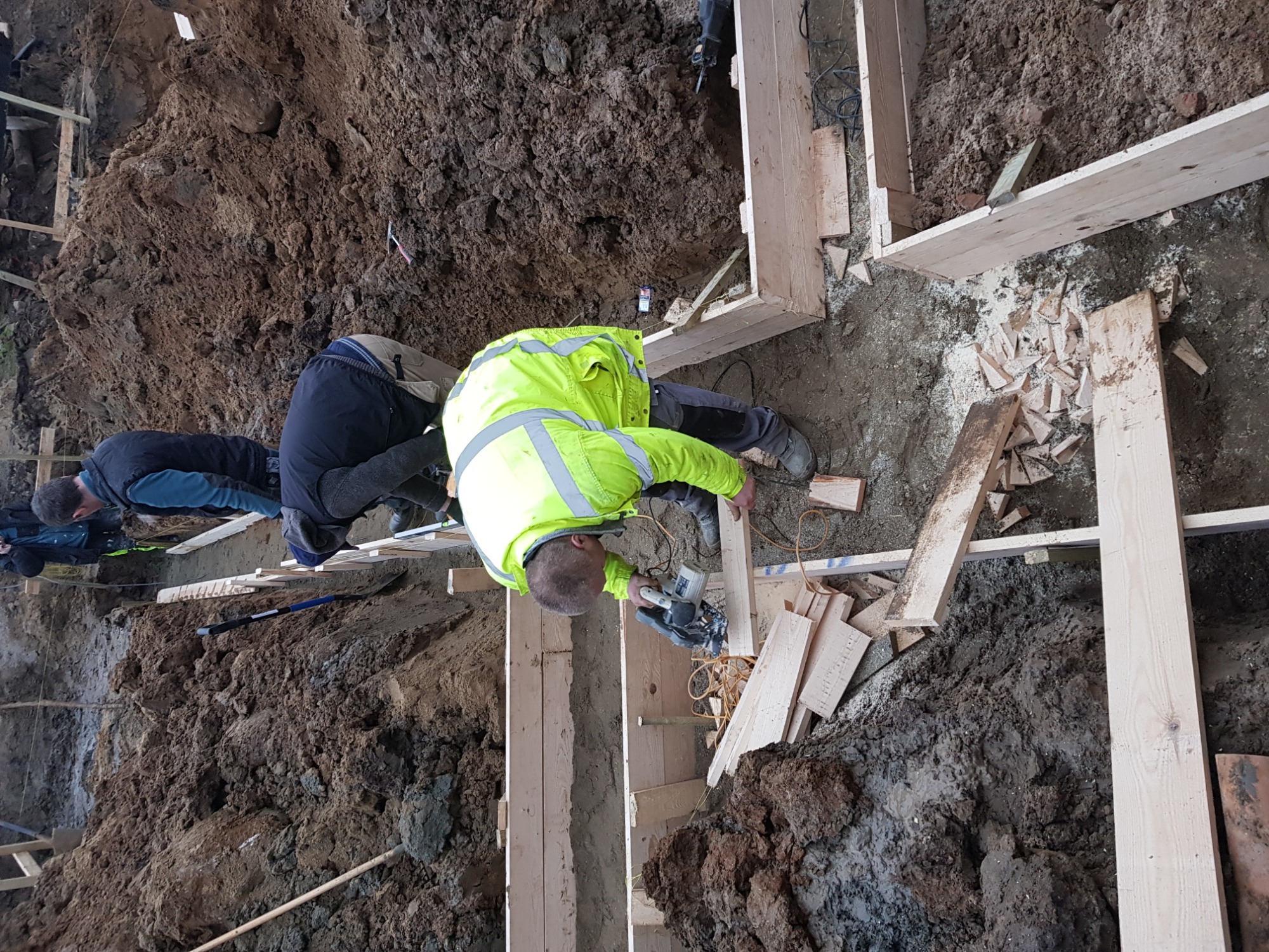 Pracownik w odblaskowej kurtce tnie drewniane deski na budowie fundamentów przy pomocy piły elektrycznej, widoczny wykop ziemny i drewniane szalunki.