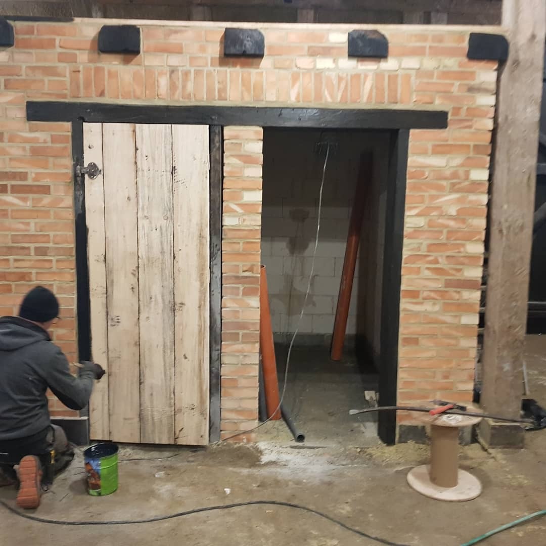 Murarz maluje na czarno ościeżnicę z surowego drewna w nowo wzniesionej ścianie z czerwonej cegły. W tle widoczne instalacje sanitarne.