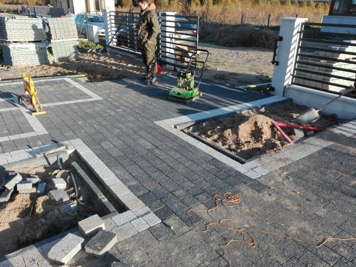 Układanie szarej kostki brukowej z jasnymi obrzeżami, częściowo ukończone, z widoczną zagęszczarką, stosem kostki, narzędziami i pracownikiem w tle.