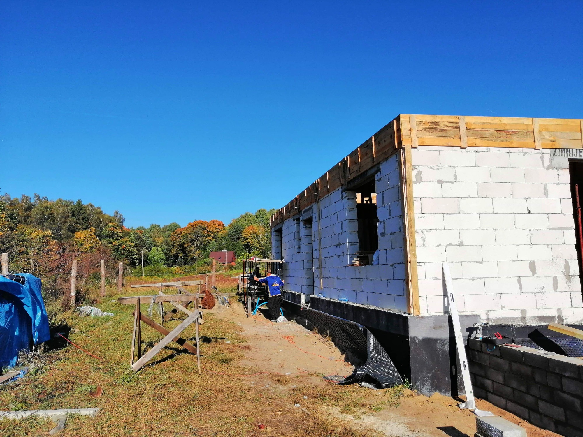 Wznoszenie ścian parterowego domu z betonu komórkowego, widoczny fundament z izolacją przeciwwilgociową i szalunek stropu, w tle drzewa i błękitne niebo.