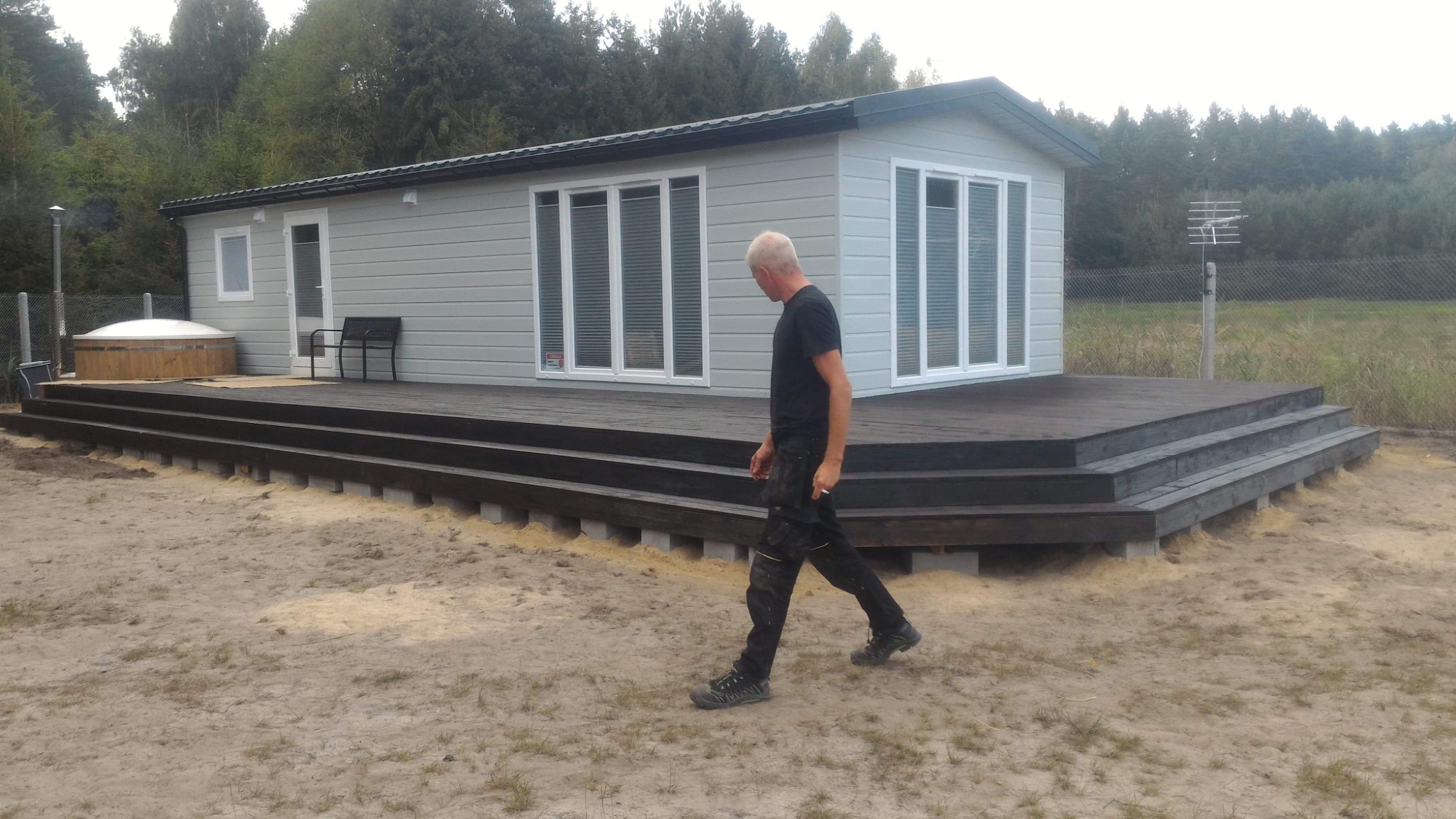 Szary dom z drewnianą elewacją i ciemnym tarasem z balustradą, widoczny mężczyzna w ubraniu roboczym na piaszczystym terenie, w tle las i antena.