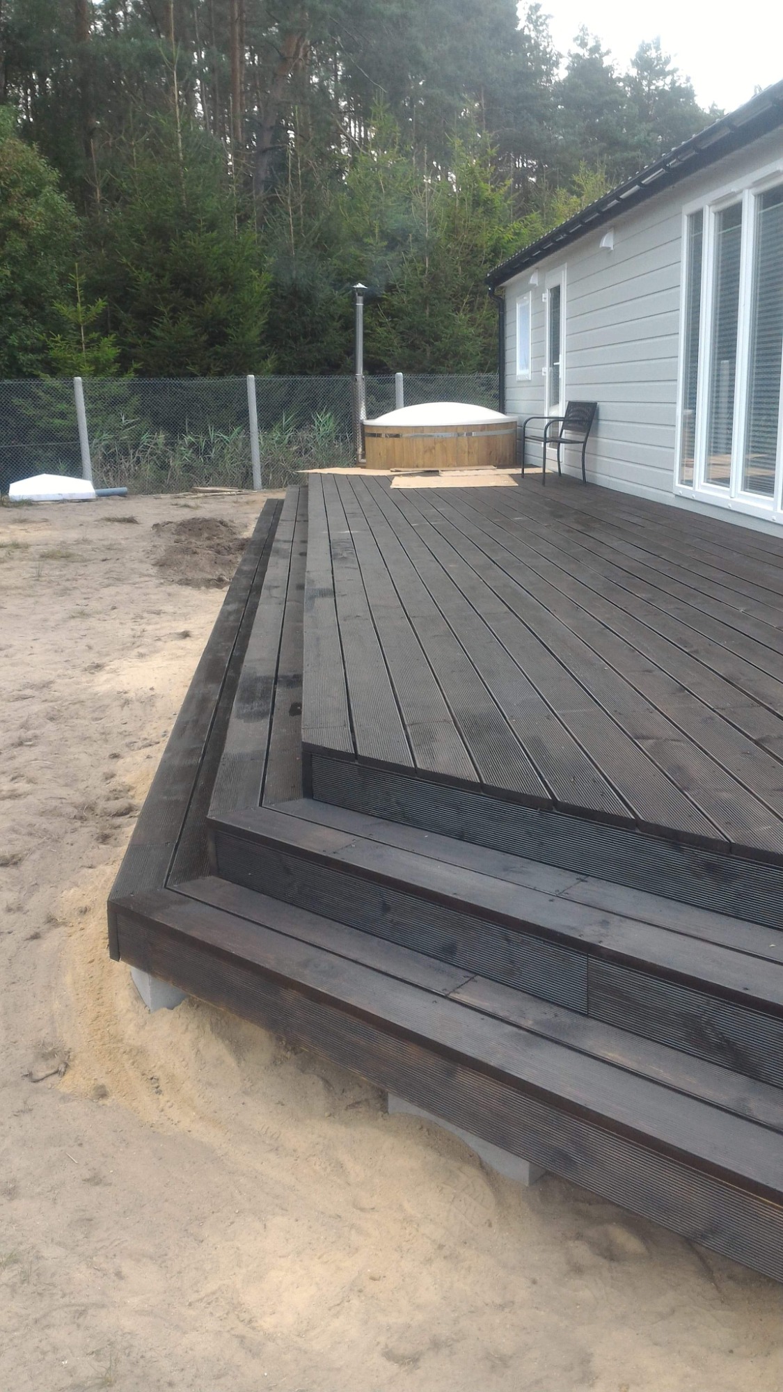Drewniany taras z schodami, obok domu z szarą elewacją i drewnianą balią z pokrywą w tle, otoczony piaszczystym terenem i drzewami.