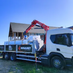 Ciężarówka z dźwigiem FASSI dostarcza białe arkusze materiału na budowę dachu domu w stanie surowym; logo Regamet na burcie.