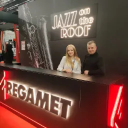 Dwie osoby za ladą recepcyjną z podświetlanym logo REGAMET na tle ściany z grafiką saksofonu i neonowym napisem 'Jazz on the Roof'.