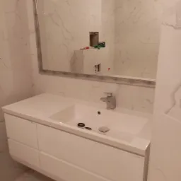 Biała umywalka z szafką i chromowaną baterią na tle marmurowych płytek ściennych z dużym lustrem w srebrnej ramie, widoczne akcesoria umywalkowe.