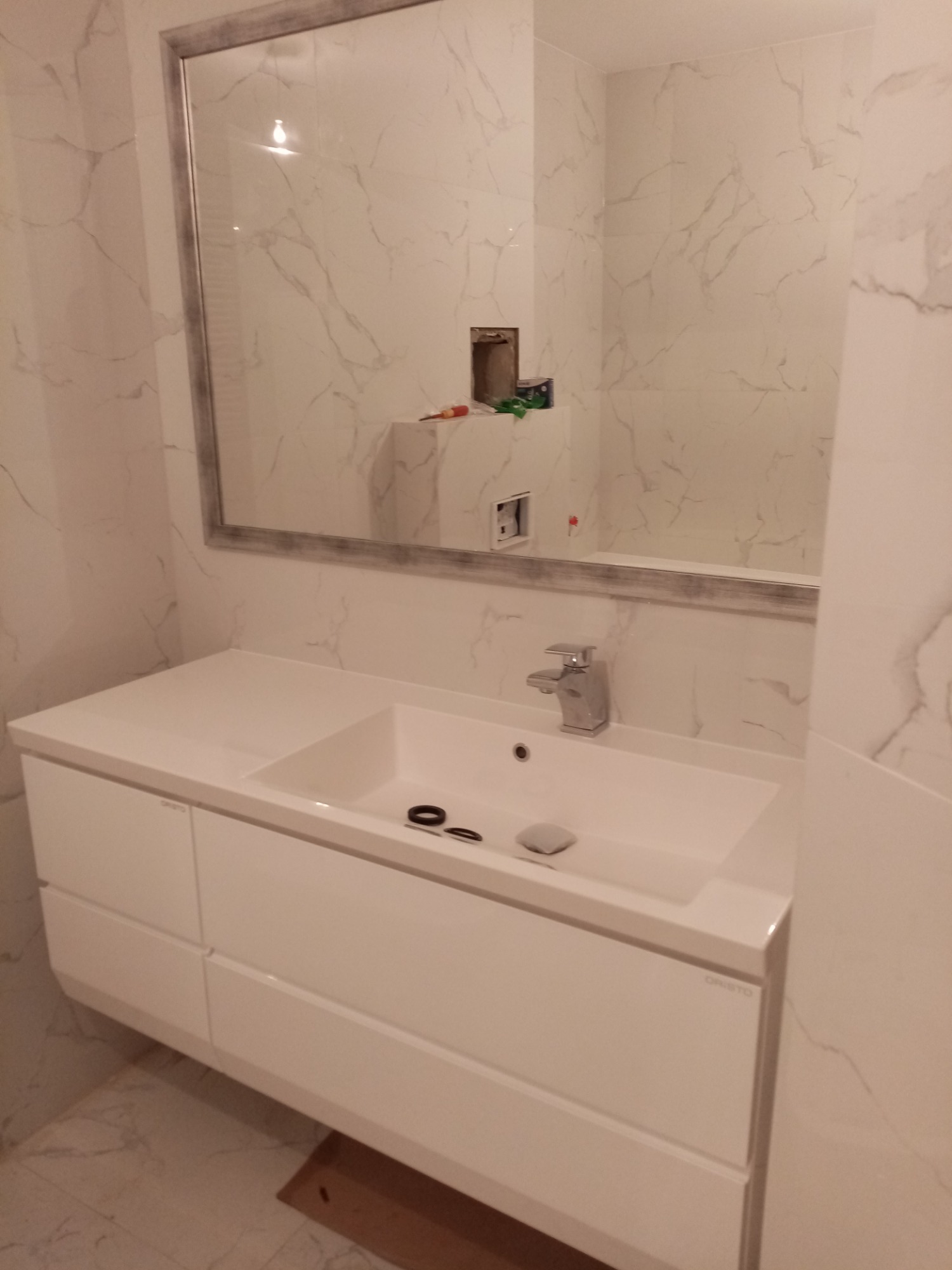 Biała umywalka z szafką i chromowaną baterią na tle marmurowych płytek ściennych z dużym lustrem w srebrnej ramie, widoczne akcesoria umywalkowe.