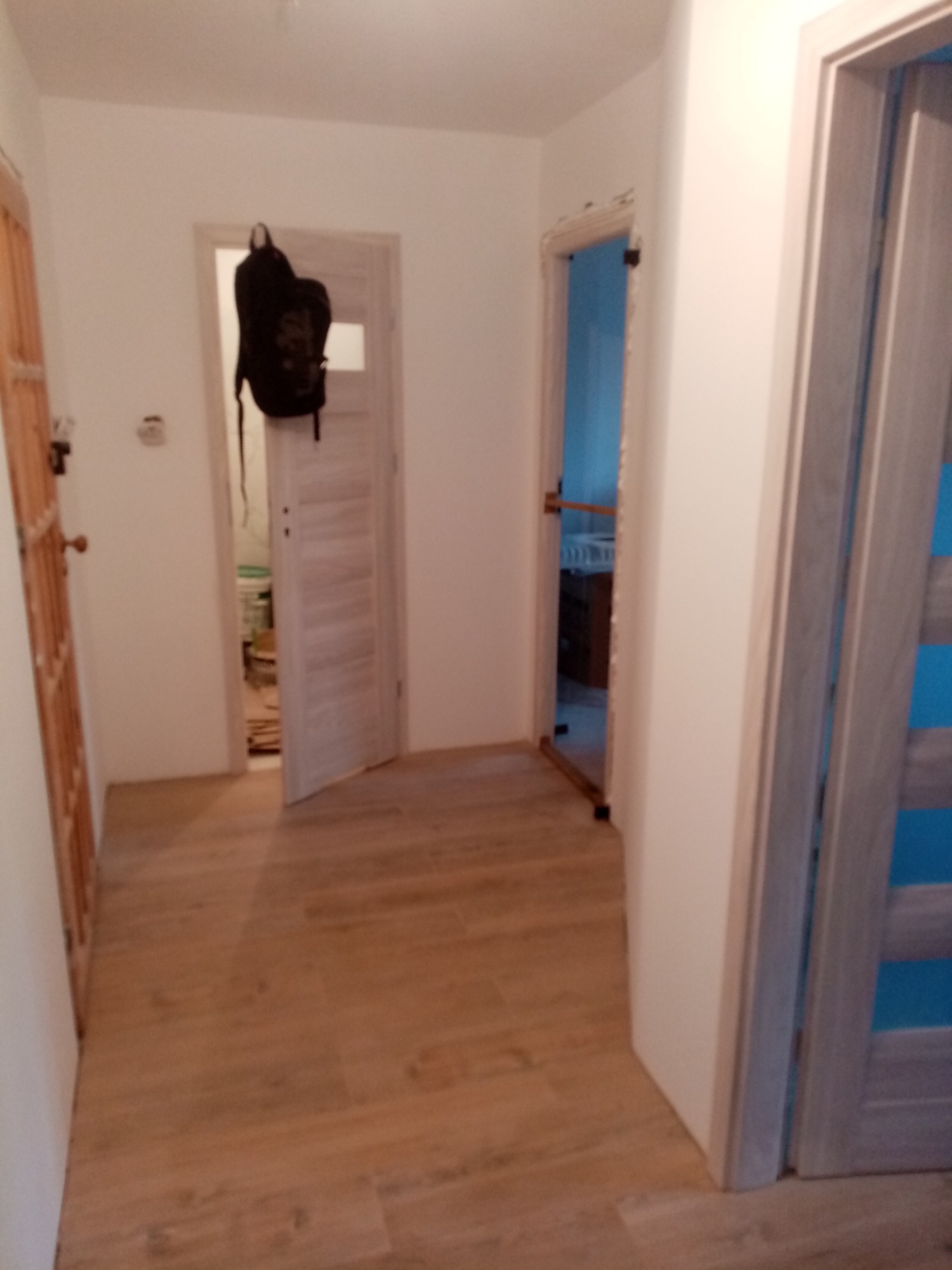 Wykończony korytarz z drewnopodobnymi płytkami podłogowymi, widoczne trzy pary drzwi w różnych odcieniach. W tle uchylone drzwi z zawieszonym czarnym plecakiem.