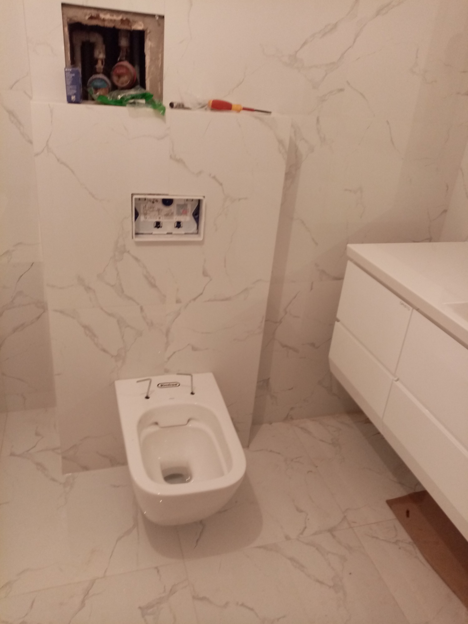 Nowoczesna, biała toaleta podwieszana w łazience z marmurowymi płytkami, widoczne elementy instalacji w ścianie, biała szafka z umywalką po prawej stronie.