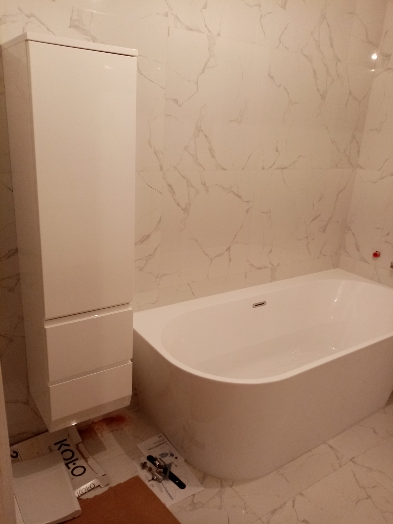 Biała, wolnostojąca wanna z odpływem, wysoka szafka łazienkowa z szufladami, marmurowe płytki ścienne i podłogowe, kartony i narzędzia montażowe na podłodze.