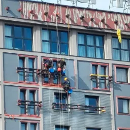 Trzech alpinistów przemysłowych zawieszonych na linach, pracujących na elewacji wysokiego budynku z szarymi panelami i ciemnymi oknami, widoczne rusztowania wiszące i zabezpieczenia.