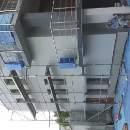 Szare, trzypiętrowe budownictwo mieszkalne w trakcie prac elewacyjnych; widoczne rusztowania, zabezpieczenia okien folią, pracownicy na rusztowaniu oraz paleta z materiałami na ziemi.