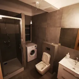 Nowoczesna łazienka z kabiną prysznicową, pralką, wiszącą toaletą z otwartą klapą i umywalką z szafką, wykończona ciemnymi płytkami, oświetlona pojedynczą żarówką na kablu.