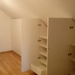 Biała szafa wnękowa z otwartymi drzwiami i wysuniętą szufladą, dopasowana do skosu poddasza, widoczna drewniana podłoga.