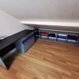 Biurko z szufladami i półki w zabudowie na poddaszu, szaro-czarno-niebieska kolorystyka, oświetlenie boczne, drewniana podłoga.