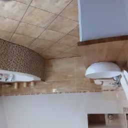 Ukośne ujęcie wykończonej łazienki z beżowymi płytkami, mozaikową obudową wanny i drewnianym blatem pod umywalką z wiszącym ręcznikiem.