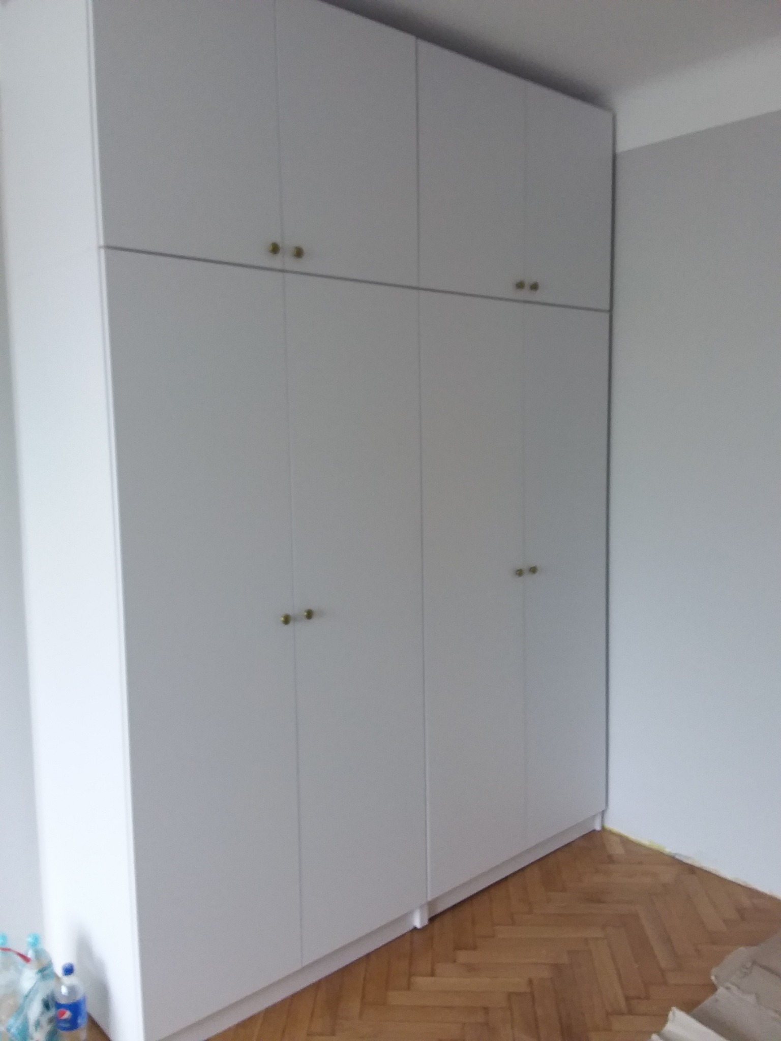 Biała, wysoka szafa z czterema drzwiami i złotymi gałkami, zamontowana w rogu pokoju w Radomyślu nad Sanem, z widoczną drewnianą podłogą w jodełkę.