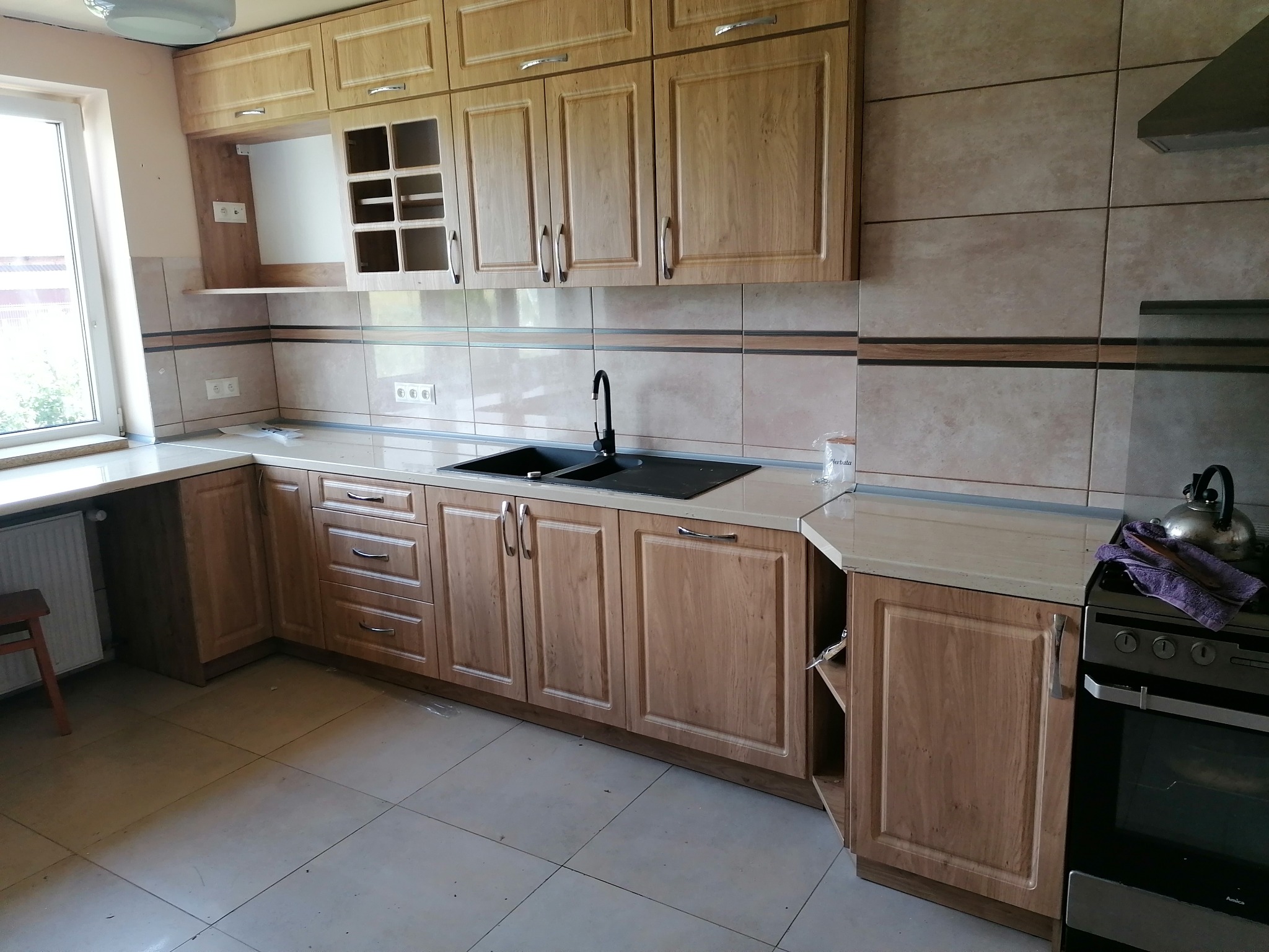 Jasna, drewniana zabudowa kuchenna z czarnym zlewem i baterią, widok z perspektywy podłogi, na blacie czajnik i fioletowa ścierka, na ścianie beżowe płytki z brązowym paskiem.