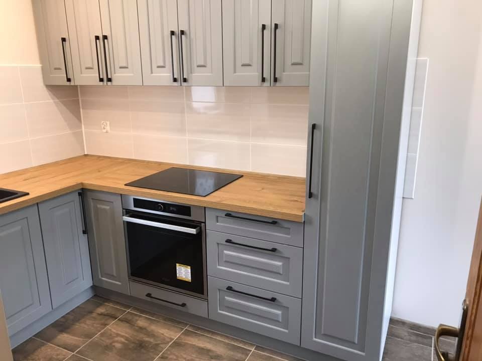 Szara kuchnia z drewnianym blatem, czarnymi uchwytami i płytą indukcyjną, widok na szafki górne i dolne oraz piekarnik w zabudowie.