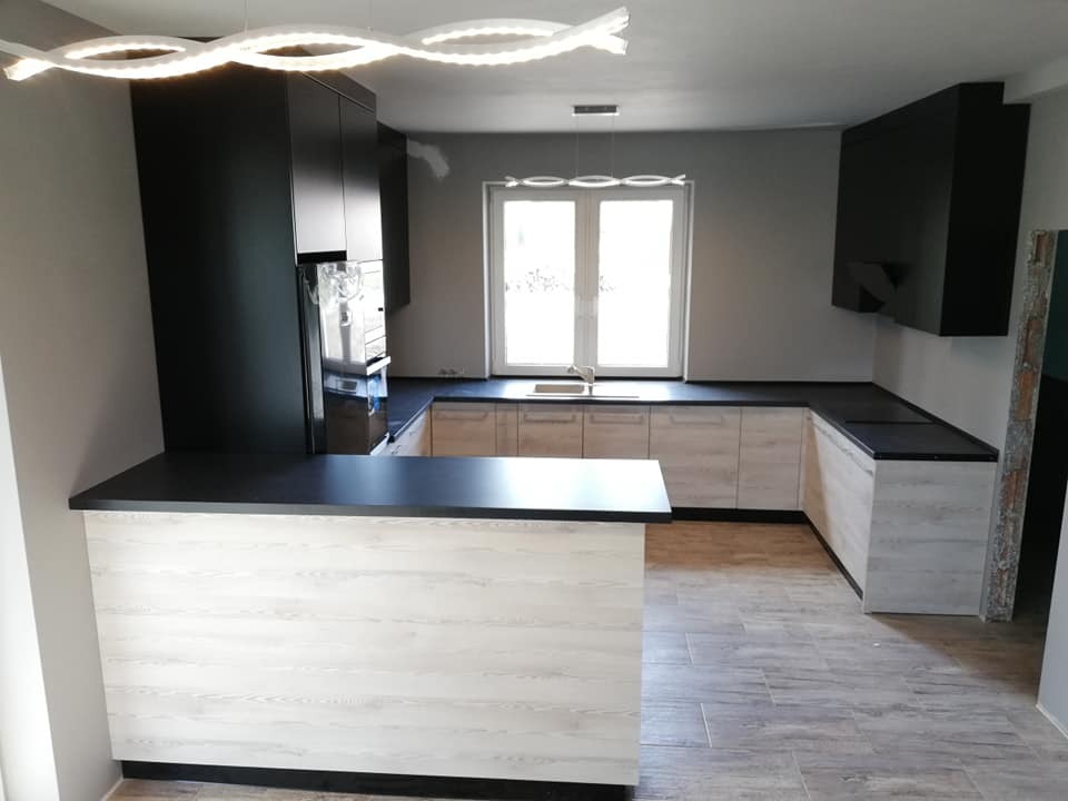 Nowoczesna kuchnia z drewnopodobnymi frontami szafek dolnych i czarnymi szafkami górnymi oraz blatem, oświetlona ledową lampą sufitową w kształcie fali.