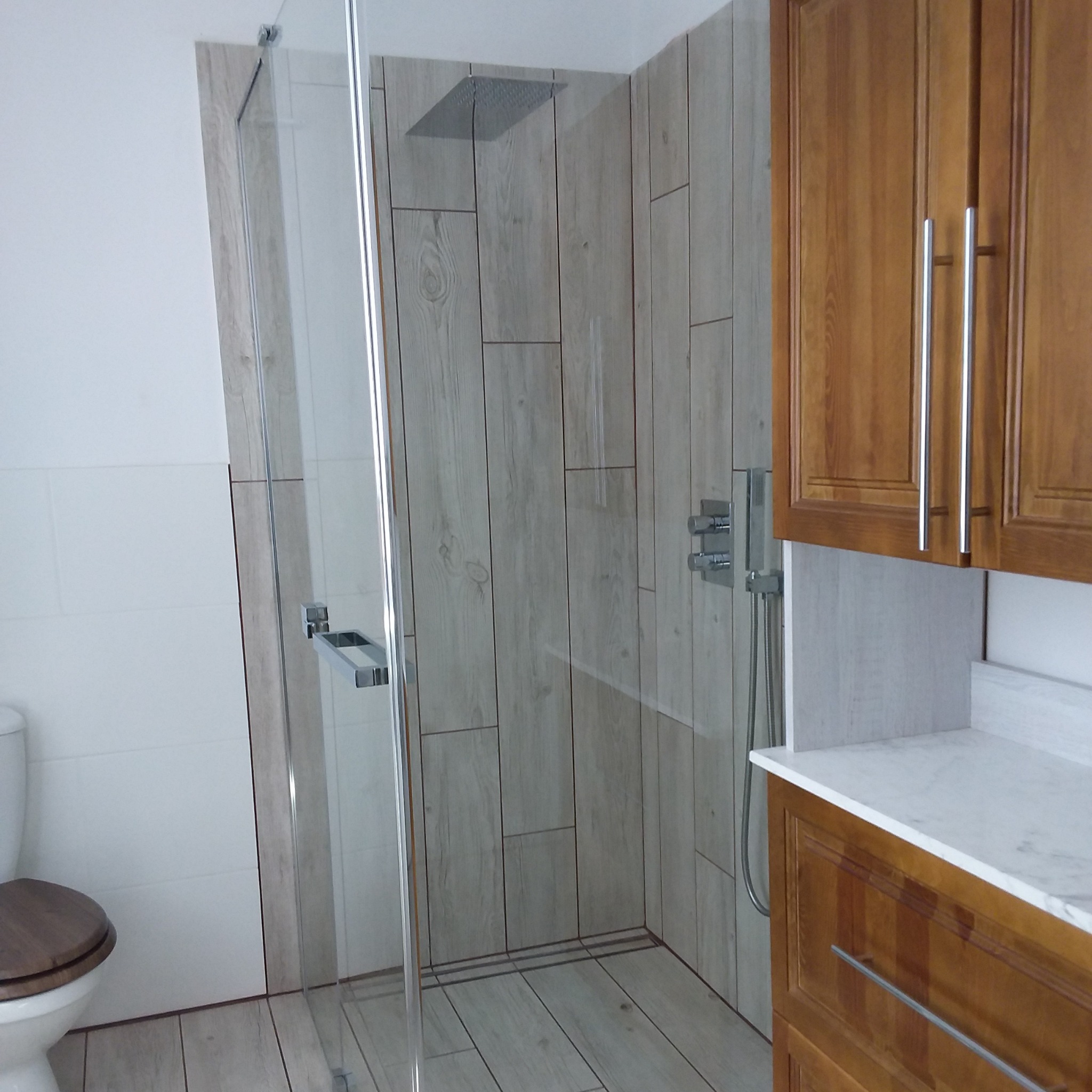 Nowoczesna kabina prysznicowa z drewnopodobnymi płytkami, deszczownicą i szklanymi drzwiami, obok szafka łazienkowa z drewnianymi frontami i marmurowym blatem.