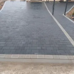 Ułożona, szara kostka brukowa na podjeździe przed domem z garażem, oddzielona jasną linią kostki, widoczne niedokończone obrzeża.