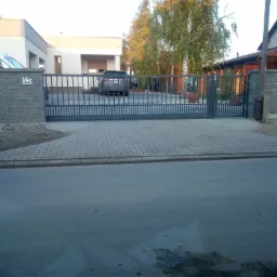 Nowoczesna, przesuwna brama wjazdowa z szarej stali, zamontowana na posesji z kostki brukowej i nowoczesnym domem w tle, widoczny samochód za bramą i tabliczka z numerem '14c' na słupku.